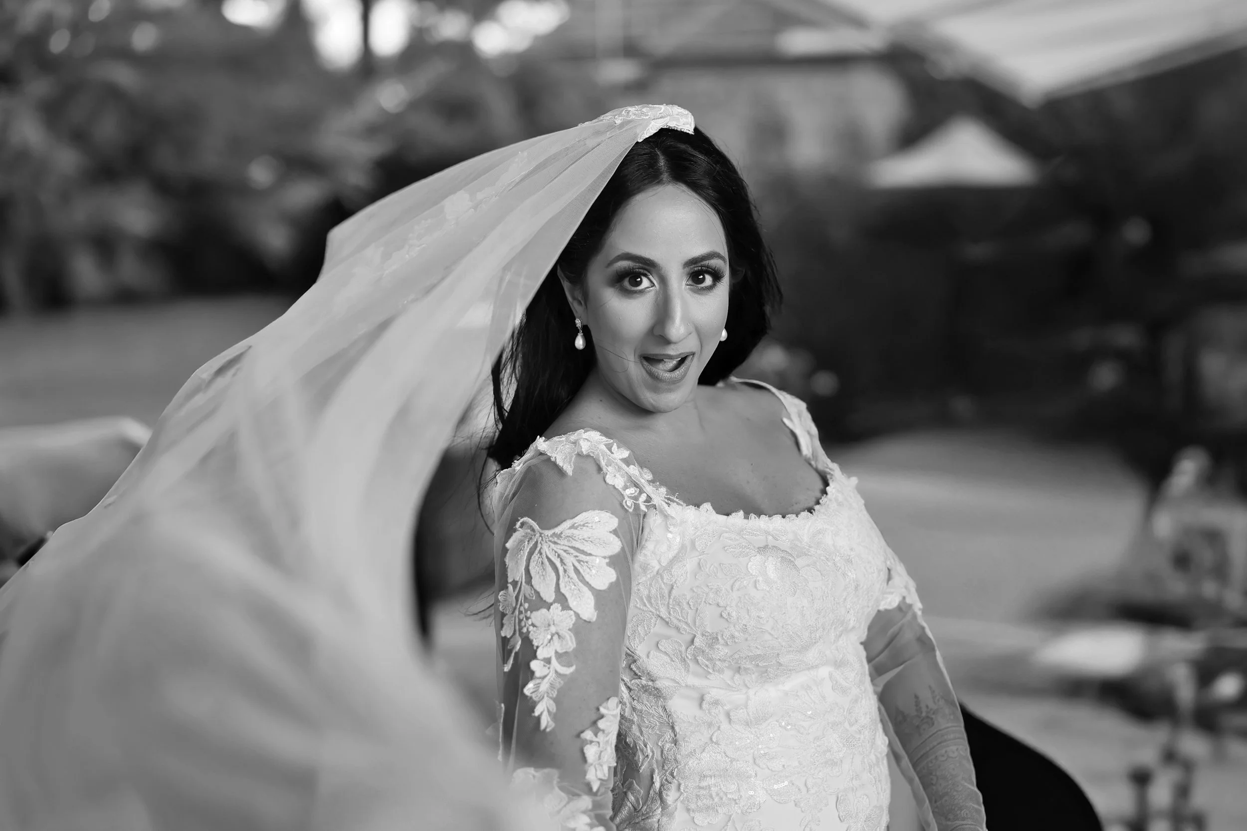 A woman dressed in a wedding gown with lace sleeves and a veil, looking surprised with her mouth slightly open, outdoors with blurred natural background.