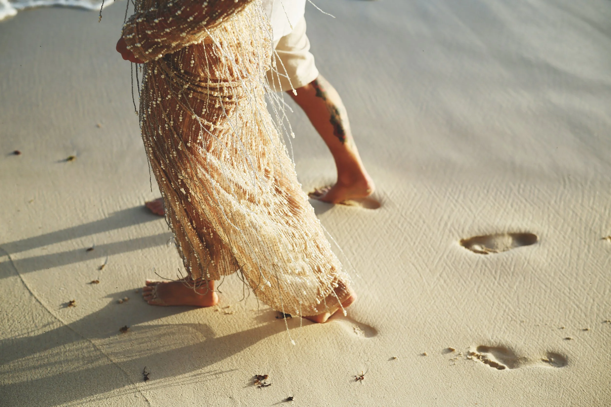 Person walking barefoot on sandy beach wearing a beige dress with intricate beadwork. Footprints visible in the sand behind them, with sunlight casting long shadows.