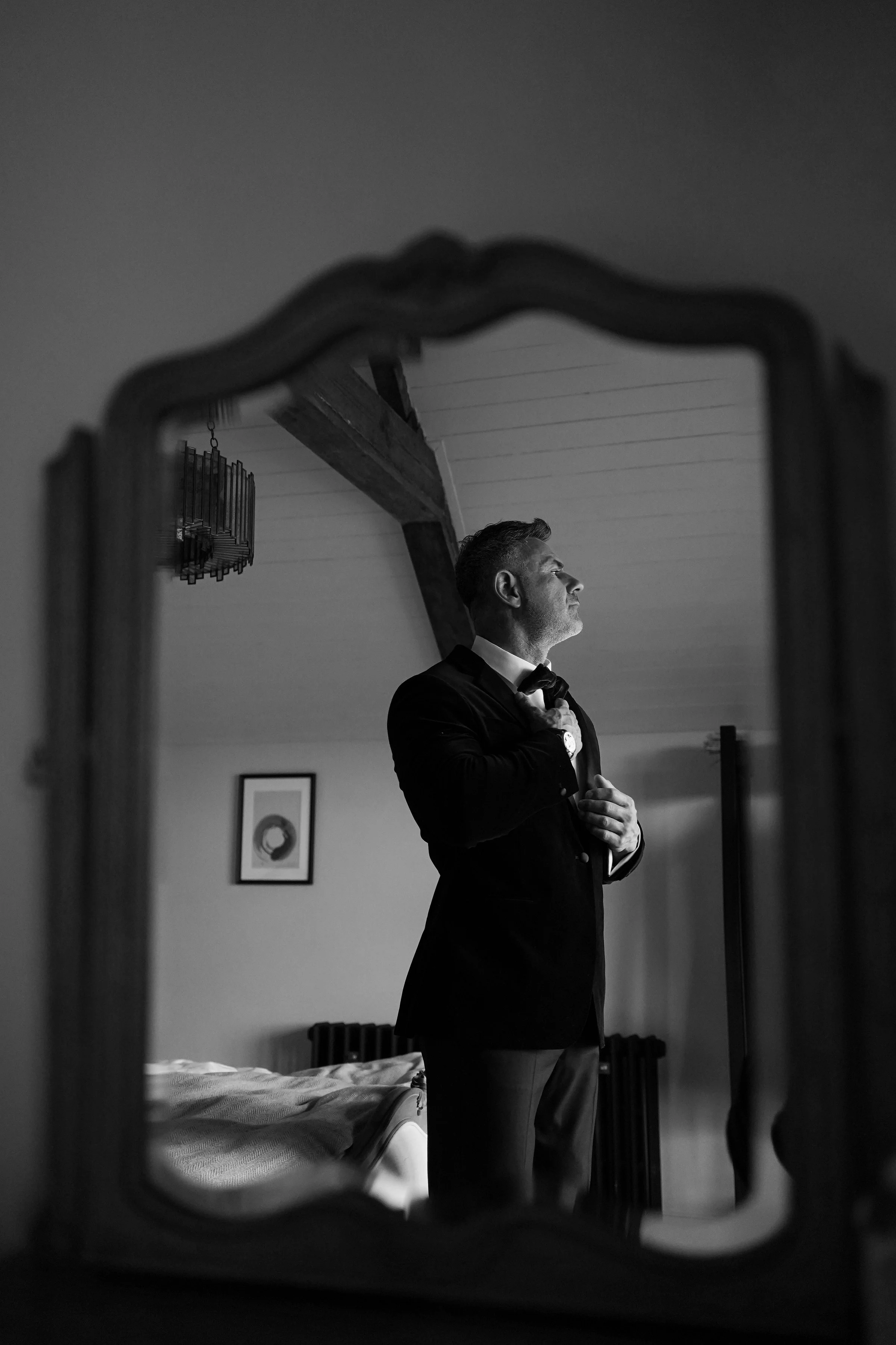 A black-and-white photo of a man getting dressed in a suit, reflected in a decorative mirror with a wavy frame. The man has short, styled hair and is adjusting his tie or collar with one hand on his chest and the other on his waist. The room has a wo