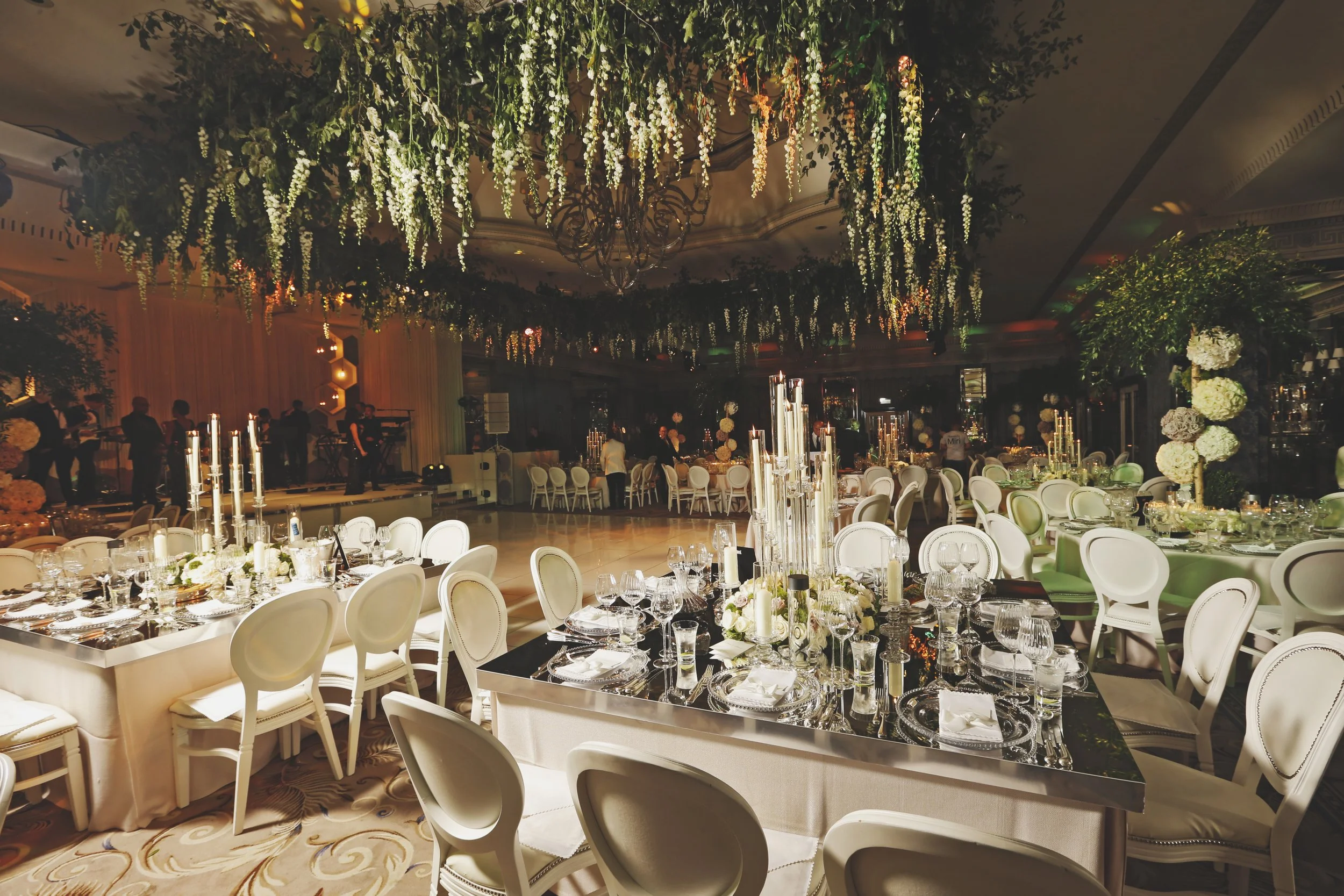 Elegant wedding reception venue with round tables, white chairs, floral centerpieces, and chandeliers. Greenery and white flowers hang from the ceiling.