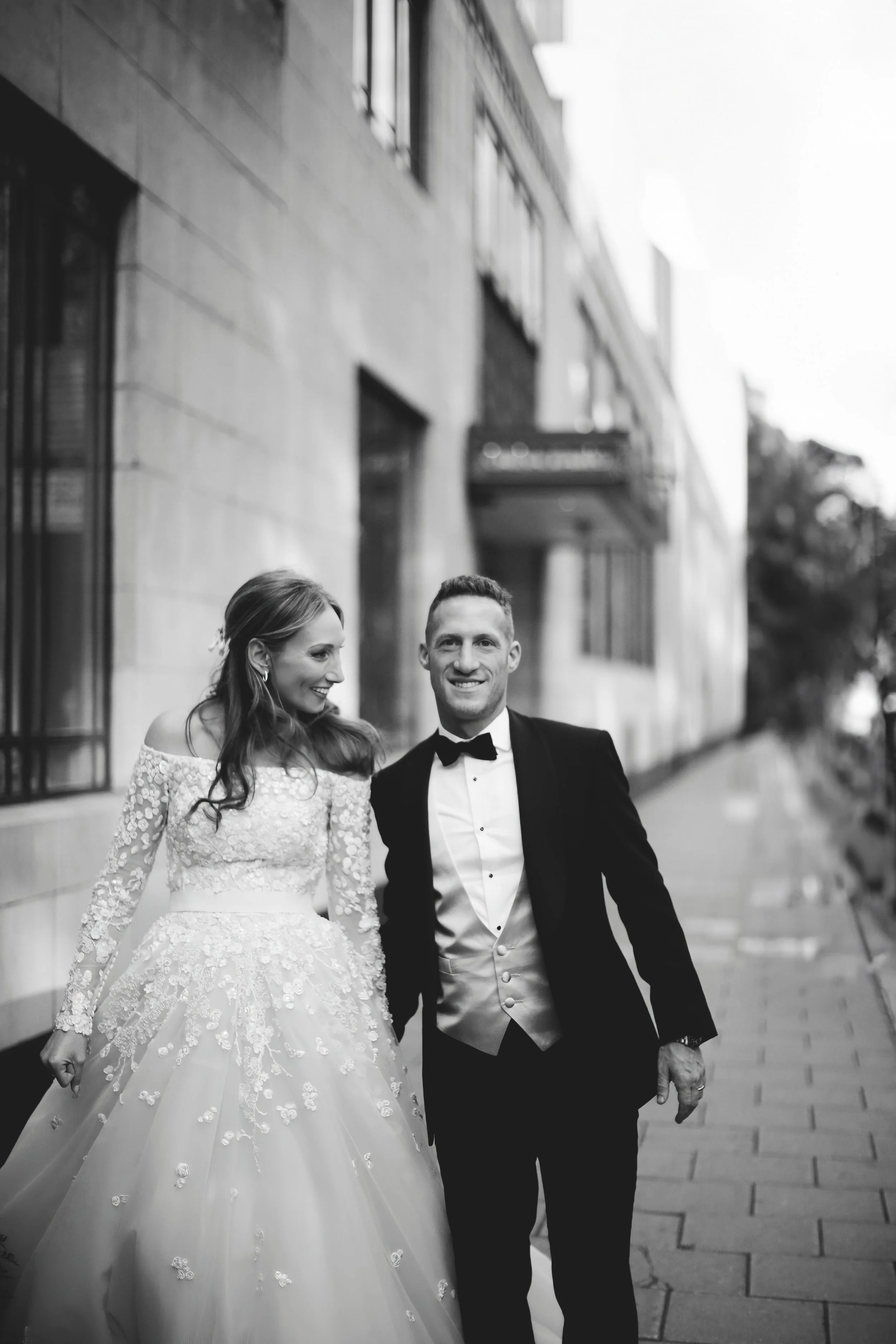 Black and white photo of a bride in a lace wedding dress and a groom in a tuxedo walking on a city sidewalk.