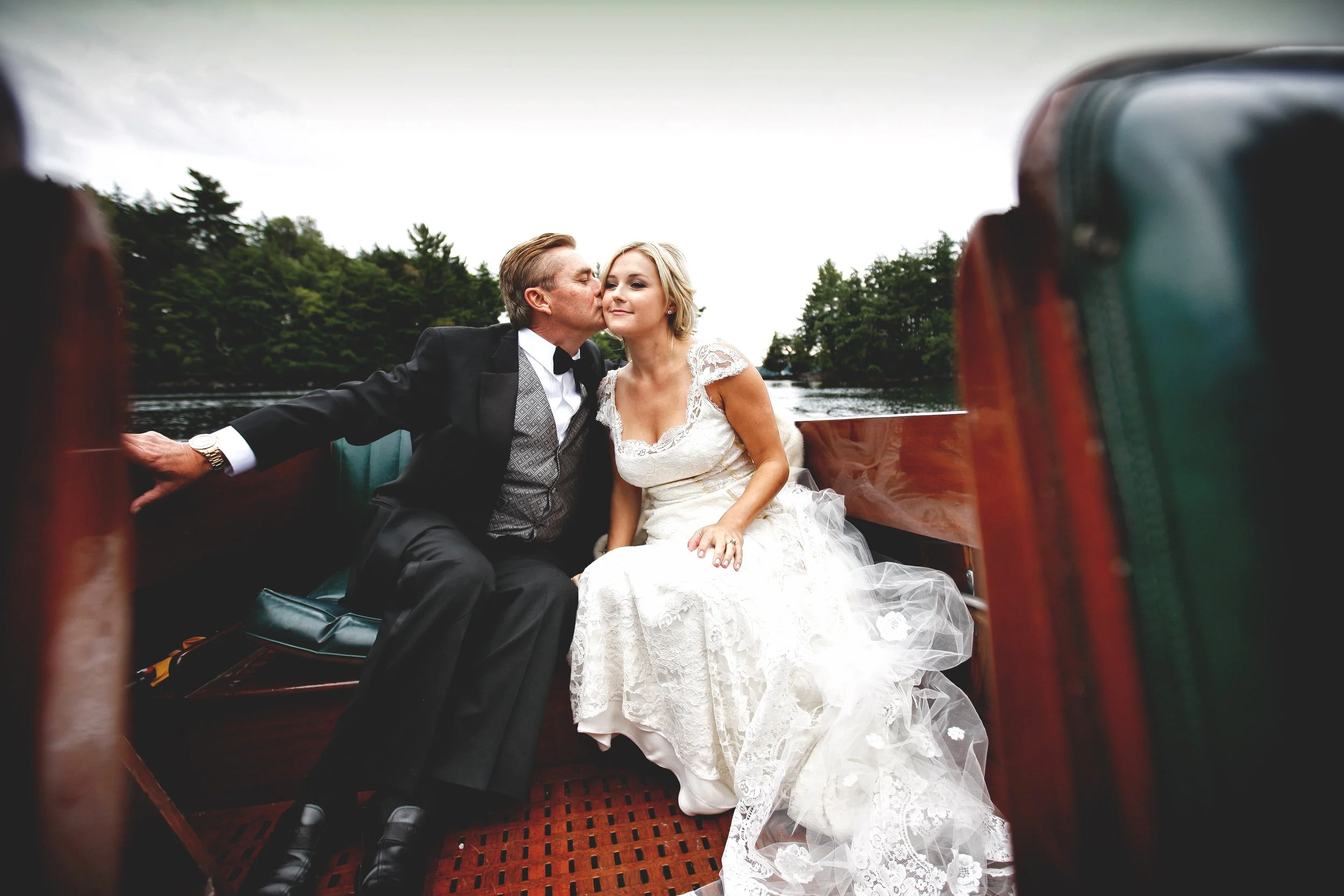 A man in a tuxedo kissing a woman in a wedding dress on a wooden boat, with trees in the background.