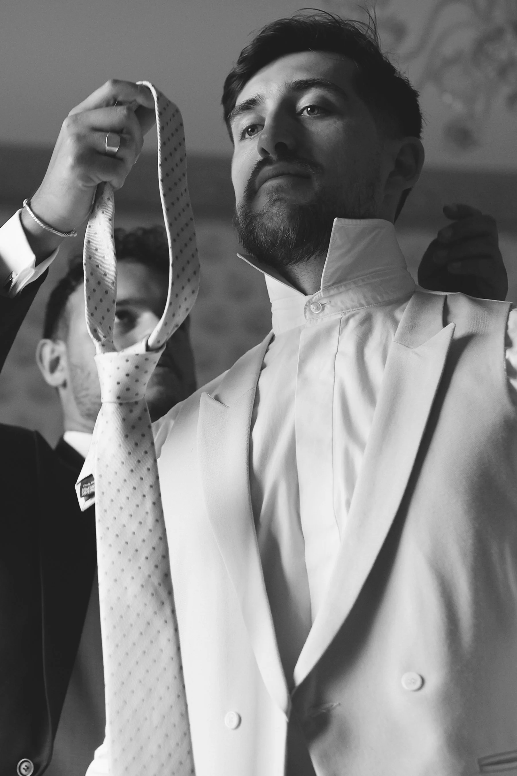 Black and white photo of a man in a white suit adjusting his tie, with another man in the background.