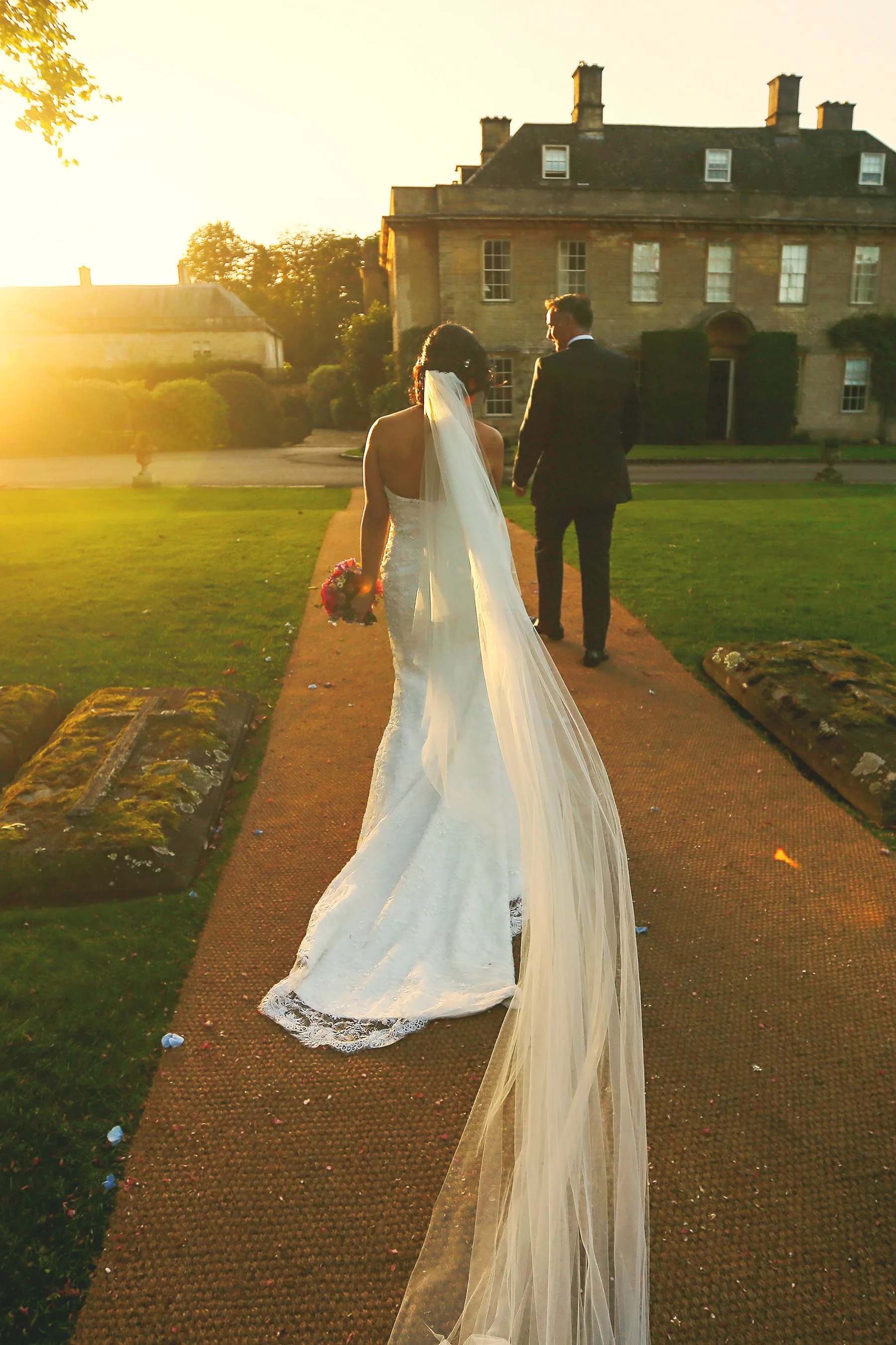 Bride and groom walking away on a churchyard path at sunset, with the bride holding a bouquet and wearing a long veil.