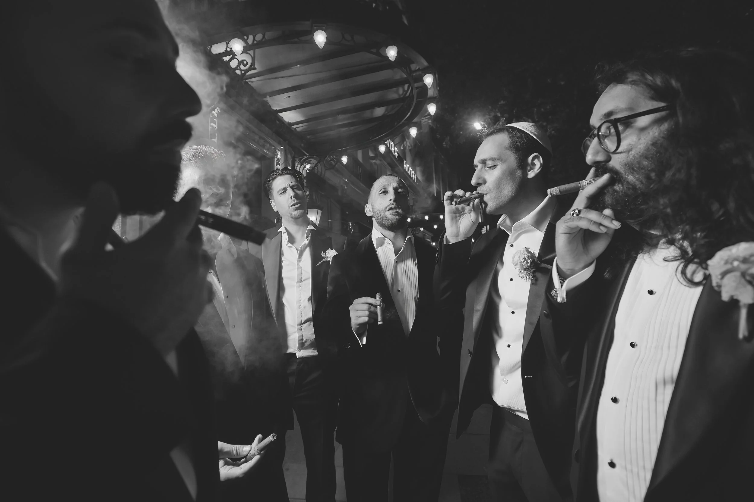 Five men in formalwear smoking cigars under canopy lighting, black and white photo.