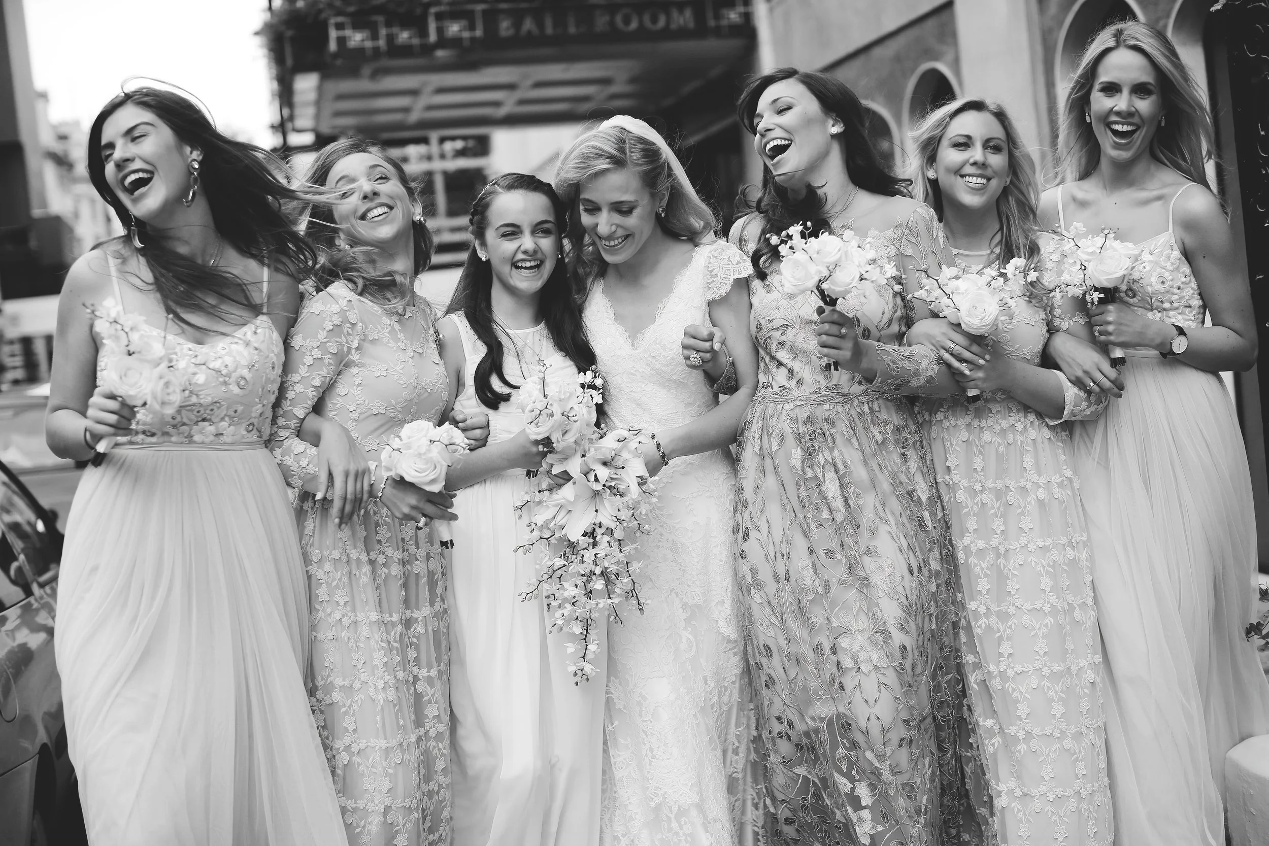 Black and white photo of a bride and bridesmaids laughing and holding bouquets, standing outdoors in elegant dresses.