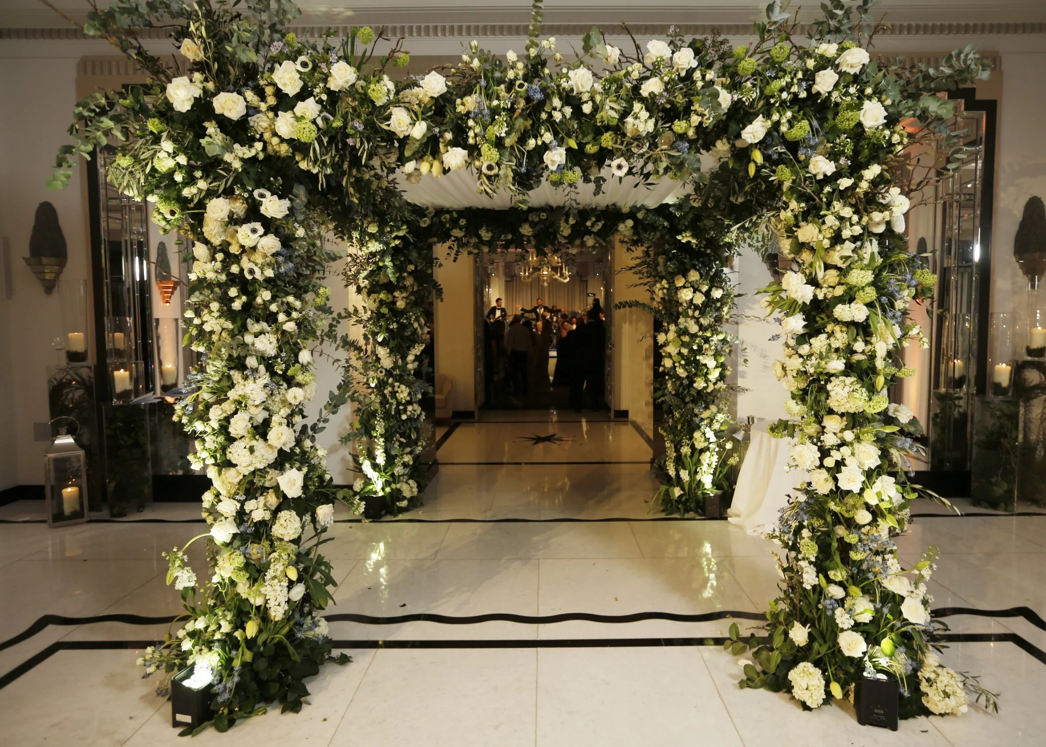 An elegant wedding arch decorated with white and green flowers and foliage at a reception hall.