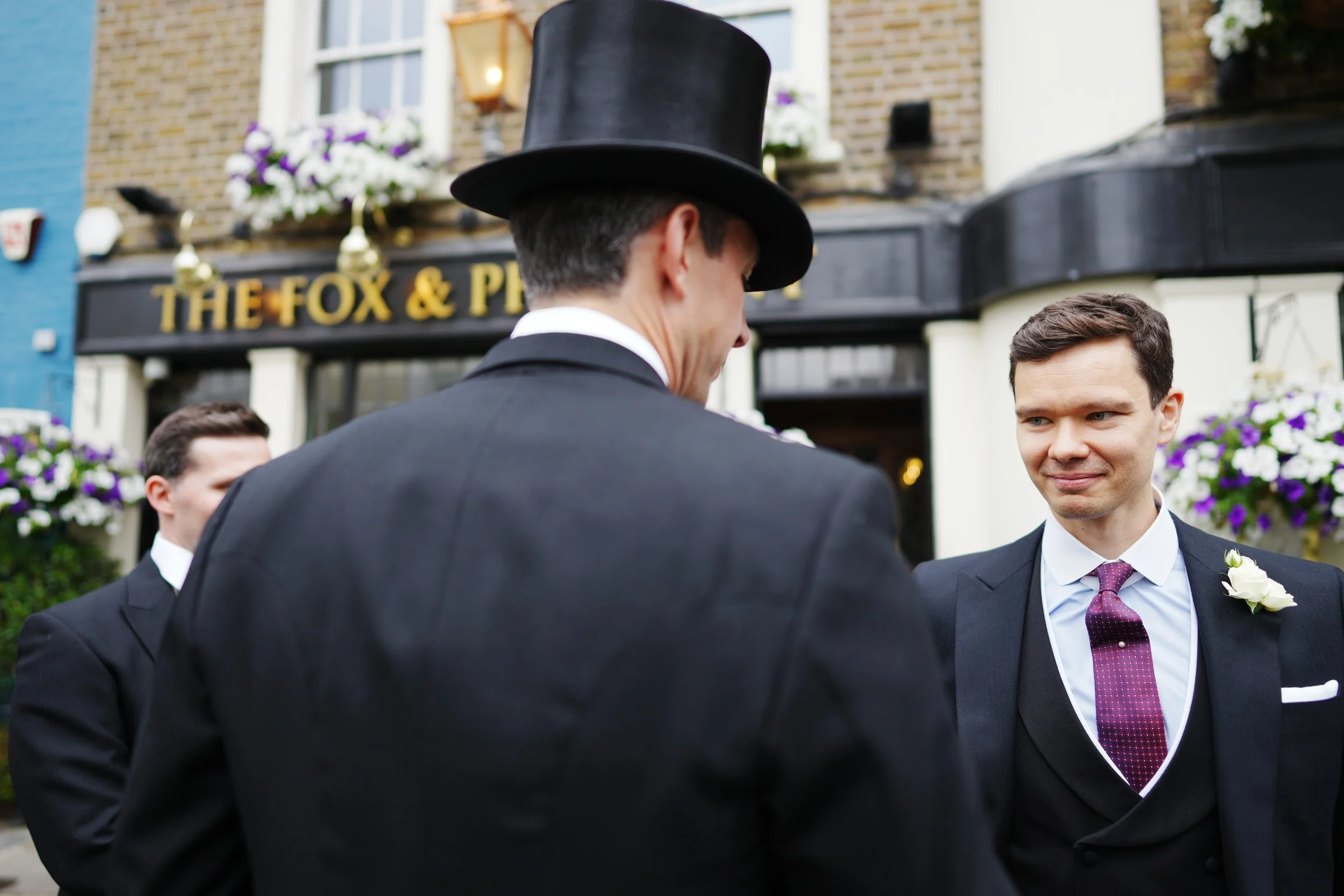 Three men dressed in formal suits and one wearing a top hat, standing outside a pub called 'The Fox & Pheasant'. One man has a boutonniere, suggesting a wedding or special occasion.