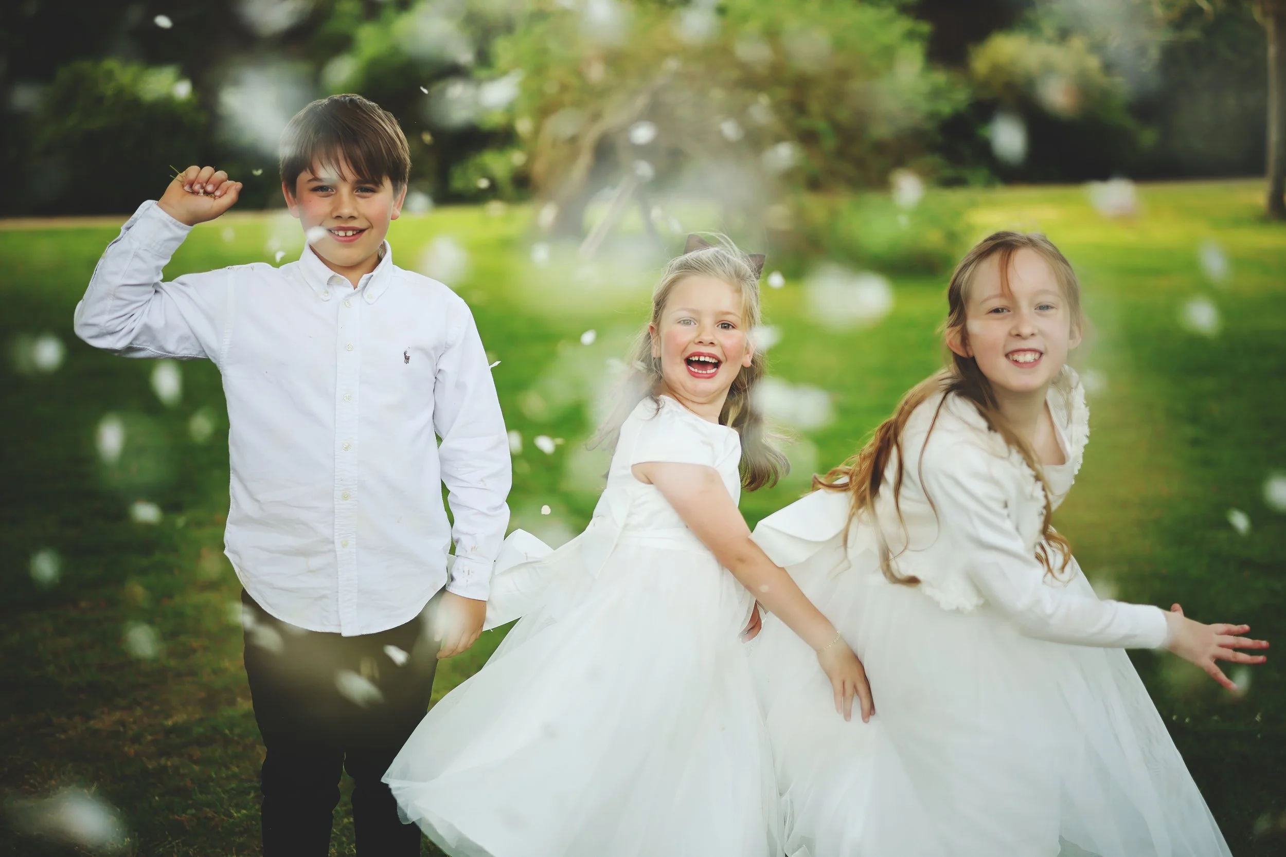 Three children, two girls and one boy, playing outdoors in a park, with the girls dressed in white dresses and the boy in a white shirt, smiling and enjoying themselves.