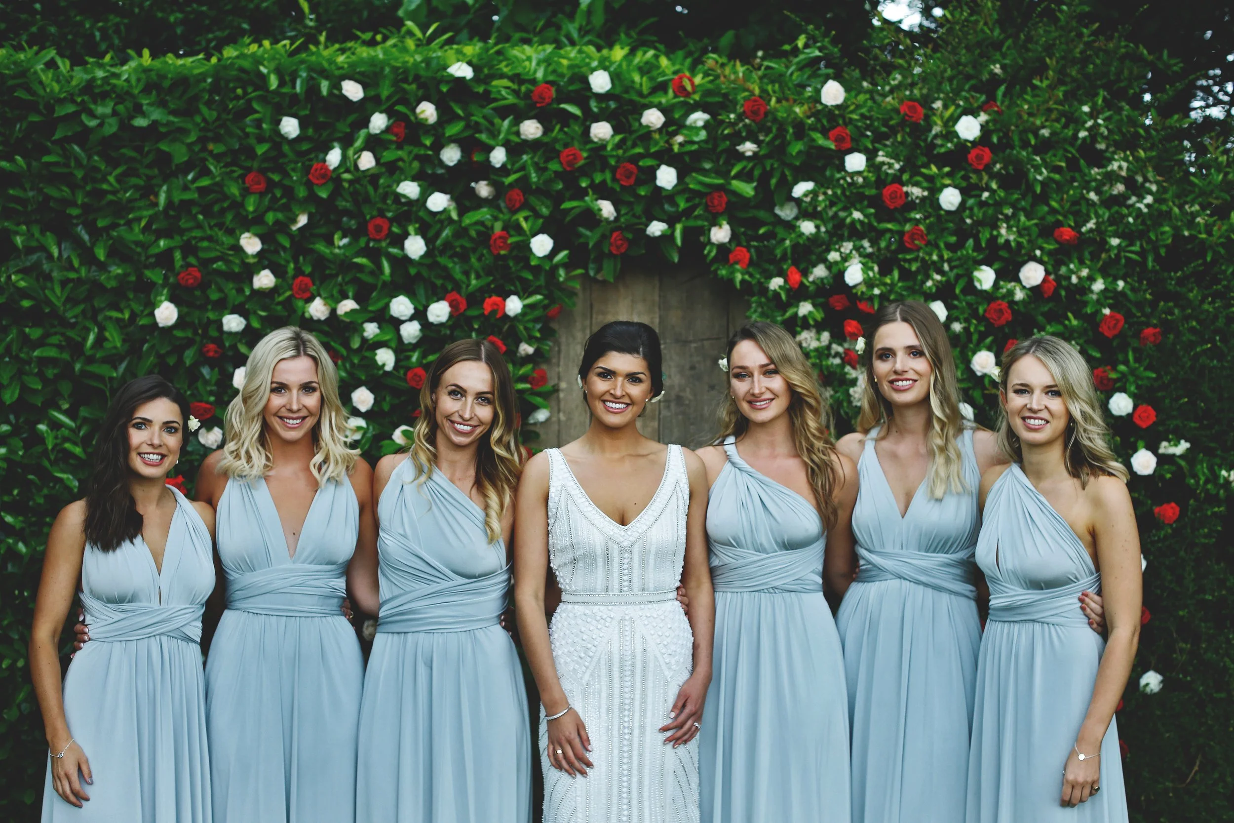 Bride and bridesmaids in light blue dresses, standing in front of a floral backdrop with red and white flowers.