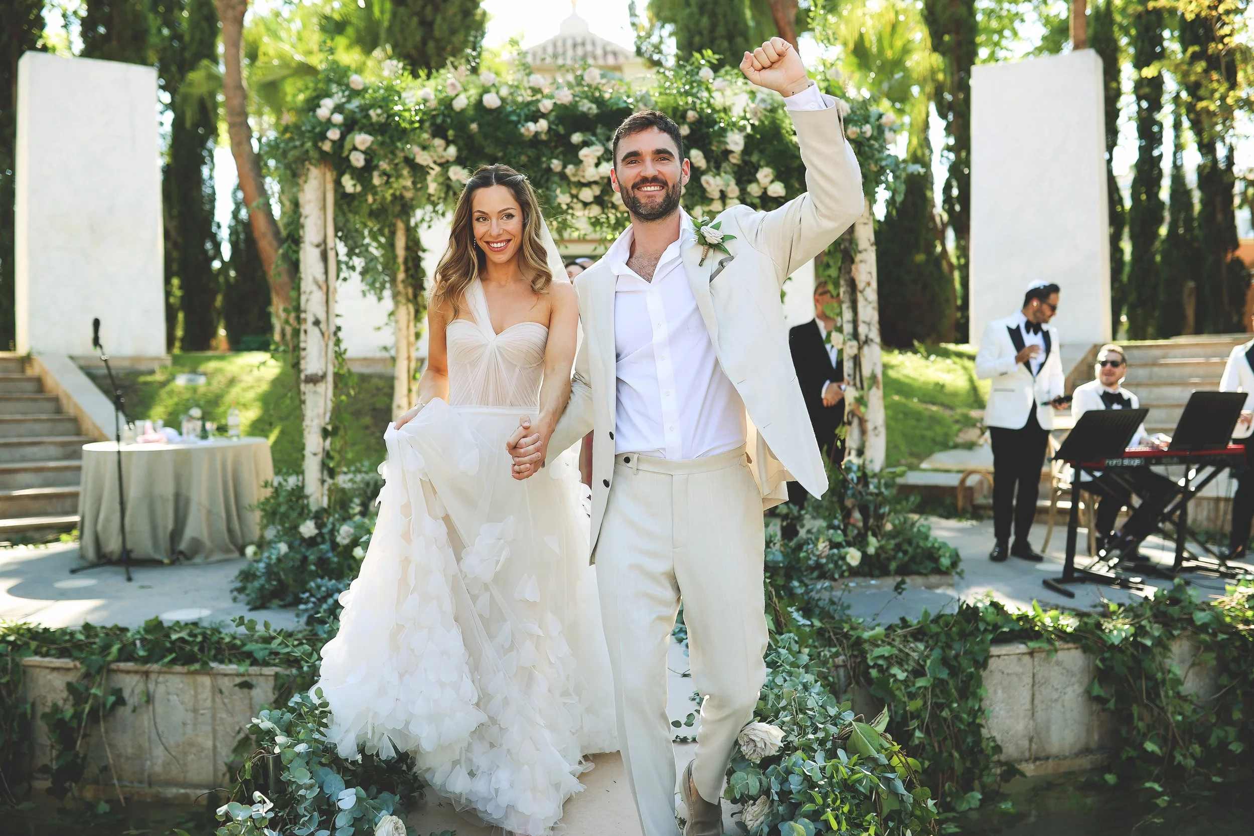 A newlywed couple celebrates after their wedding ceremony, standing under a floral arch. The groom is in a light-colored suit and the bride in a flowing white dress. Musicians in formal attire are visible in the background.