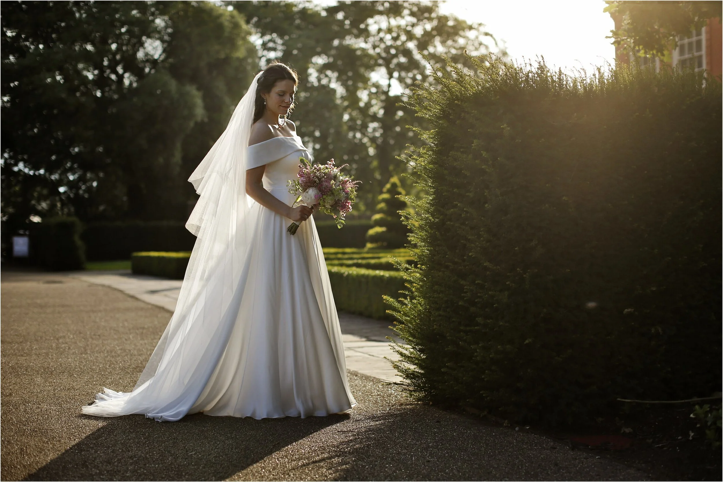 A bride in a white wedding gown holding a bouquet of pink, white, and purple flowers, standing outdoors during sunset near a tall hedge.