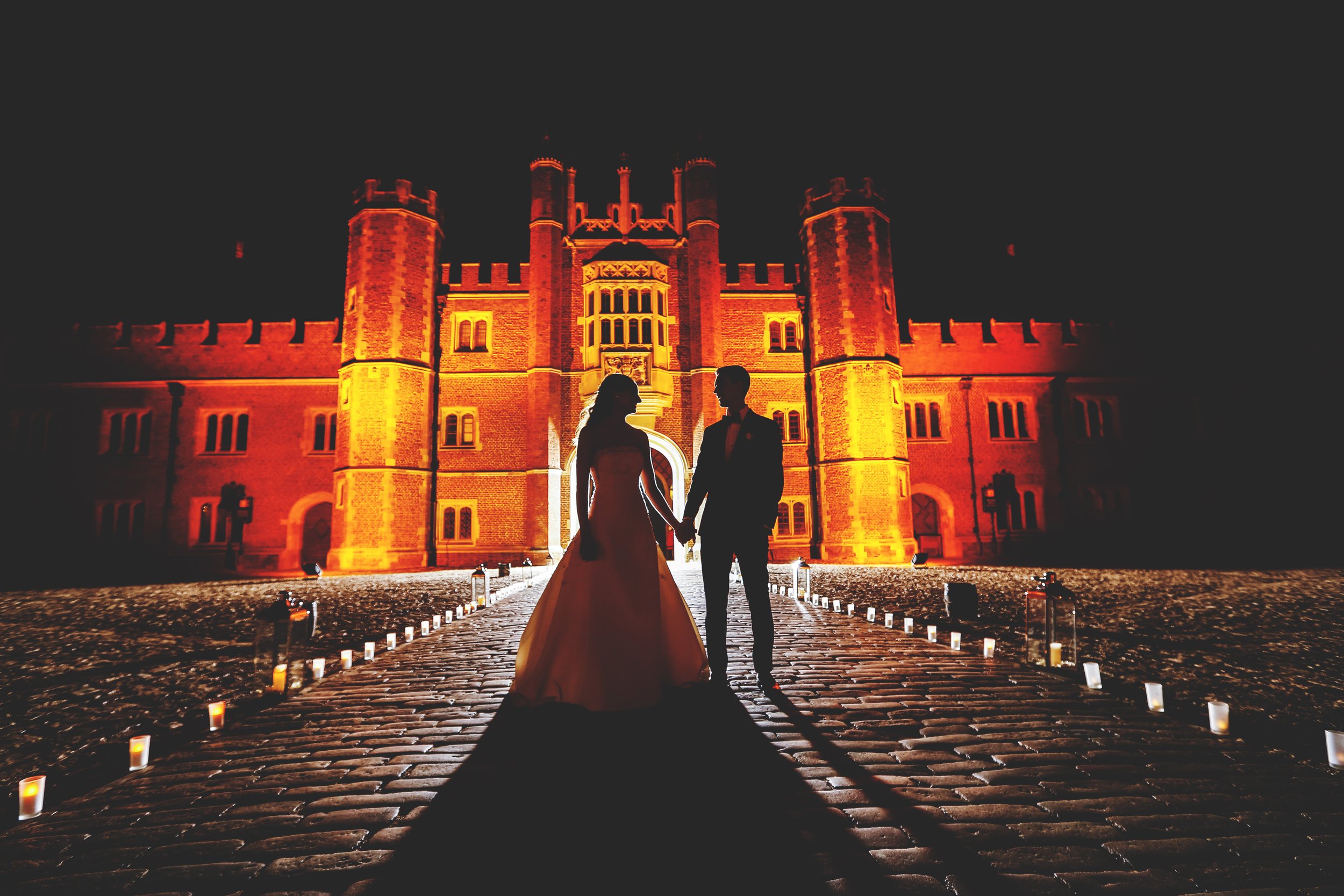 Silhouetted couple holding hands in front of a brightly lit castle at night.