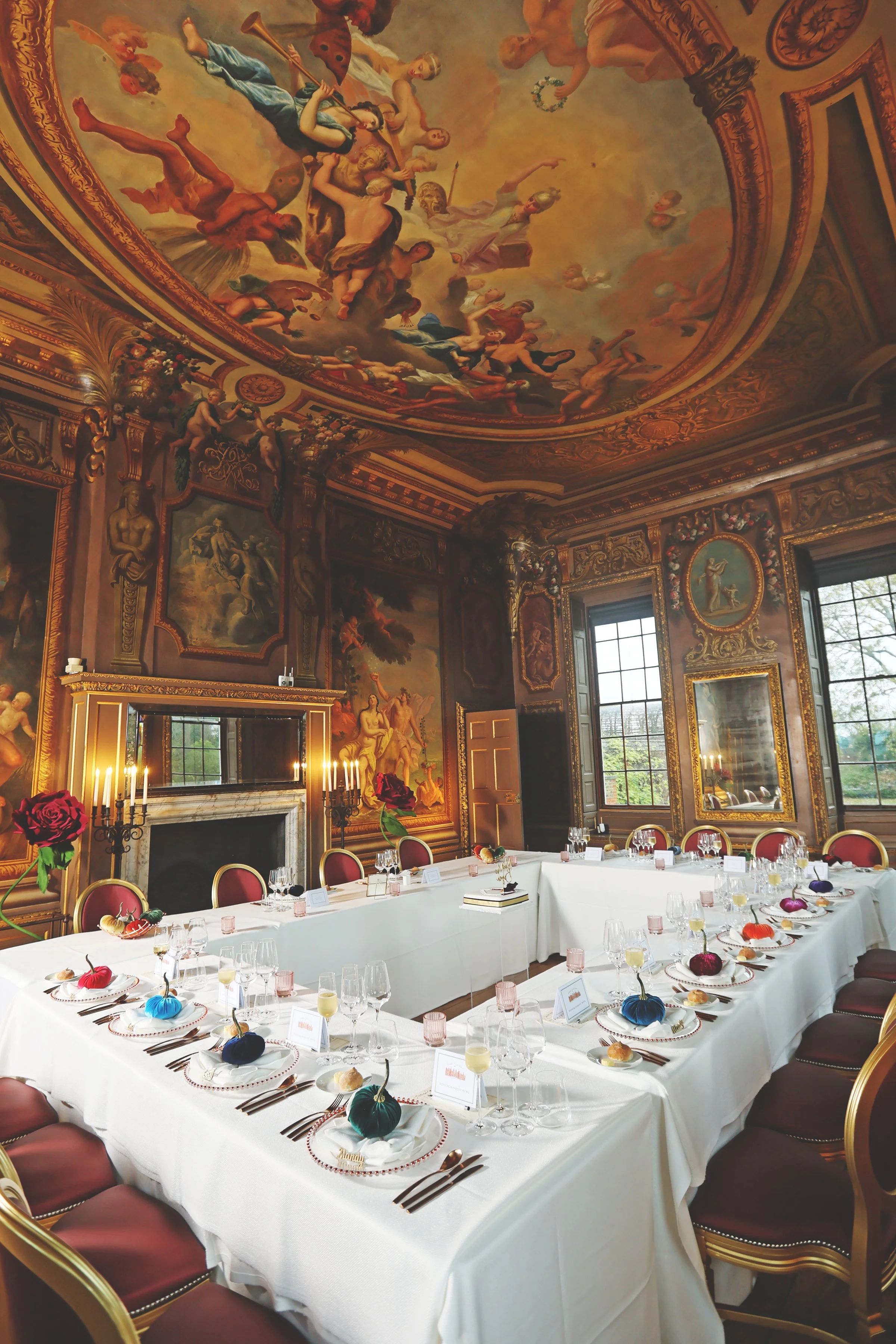Elegant dining room with ornate ceiling and walls, featuring classical paintings and intricate moldings. Tables are set with white tablecloths, colorful decorative items, plates, glassware, and candlesticks. Red chairs surround the tables.
