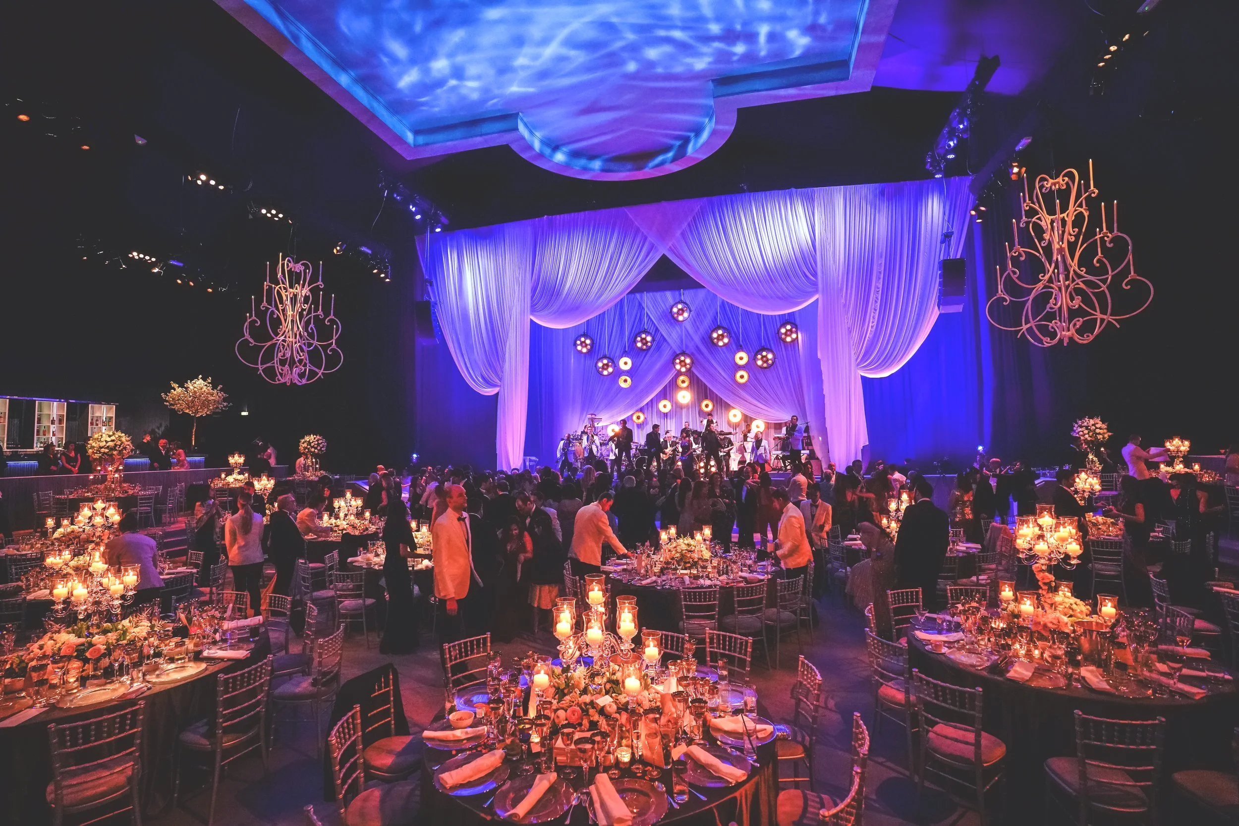 Elegant banquet hall with tables set for a formal event, chandeliers, stage with performers, and guests mingling.