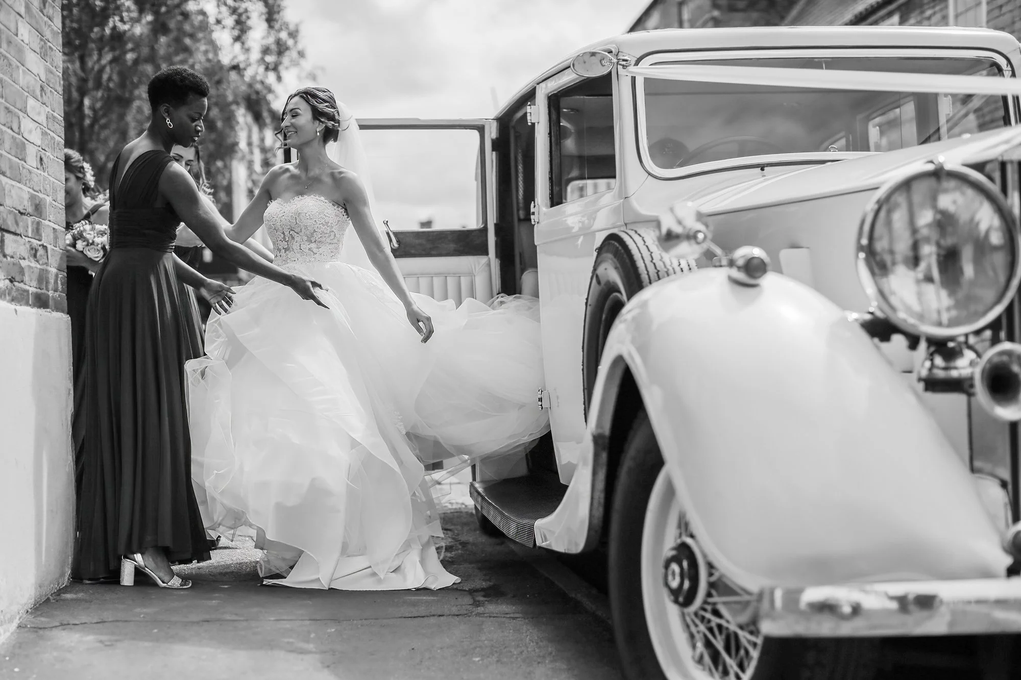 Black and white photo of a bride in a wedding dress and bridesmaid in a dress, standing next to a vintage car, smiling and holding hands outdoors.