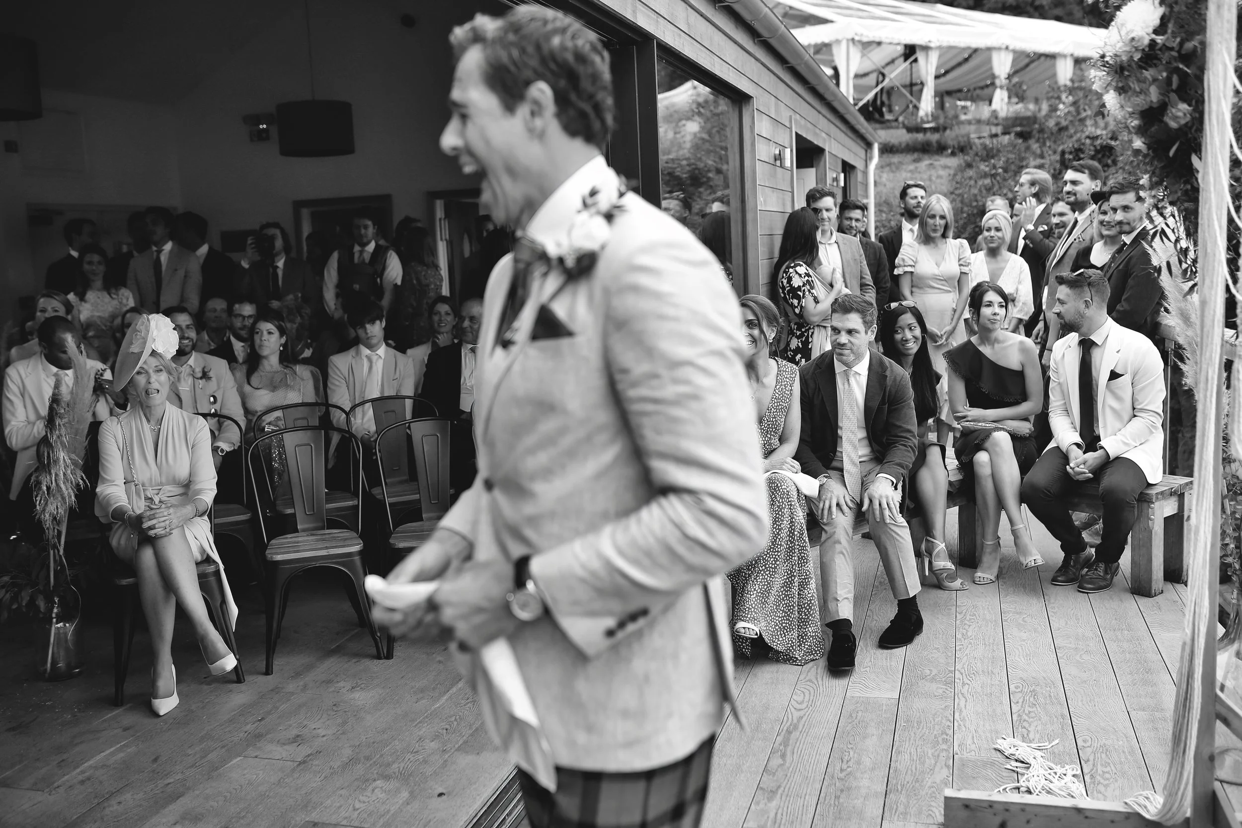 Wedding ceremony with guests seated and smiling, man in light-colored suit in foreground, indoor and outdoor sections visible.