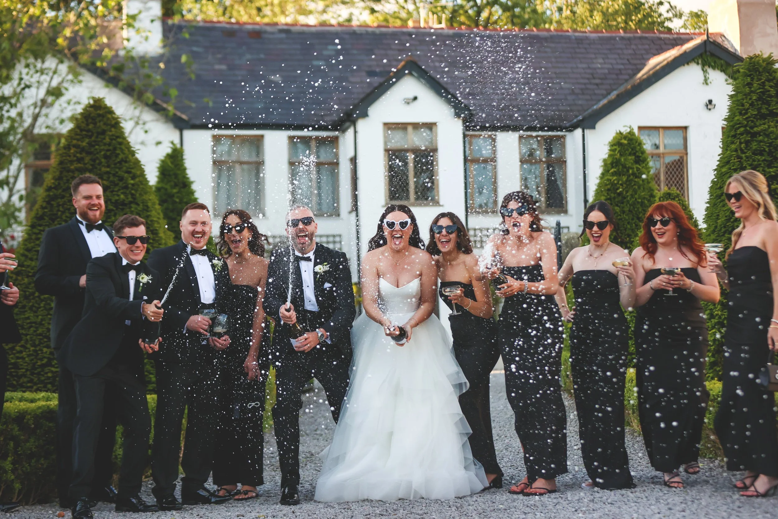 Happy wedding party celebrating outside of a house, with champagne bottles spraying confetti, with the bride in a white wedding dress and the guests in black dresses and tuxedos.