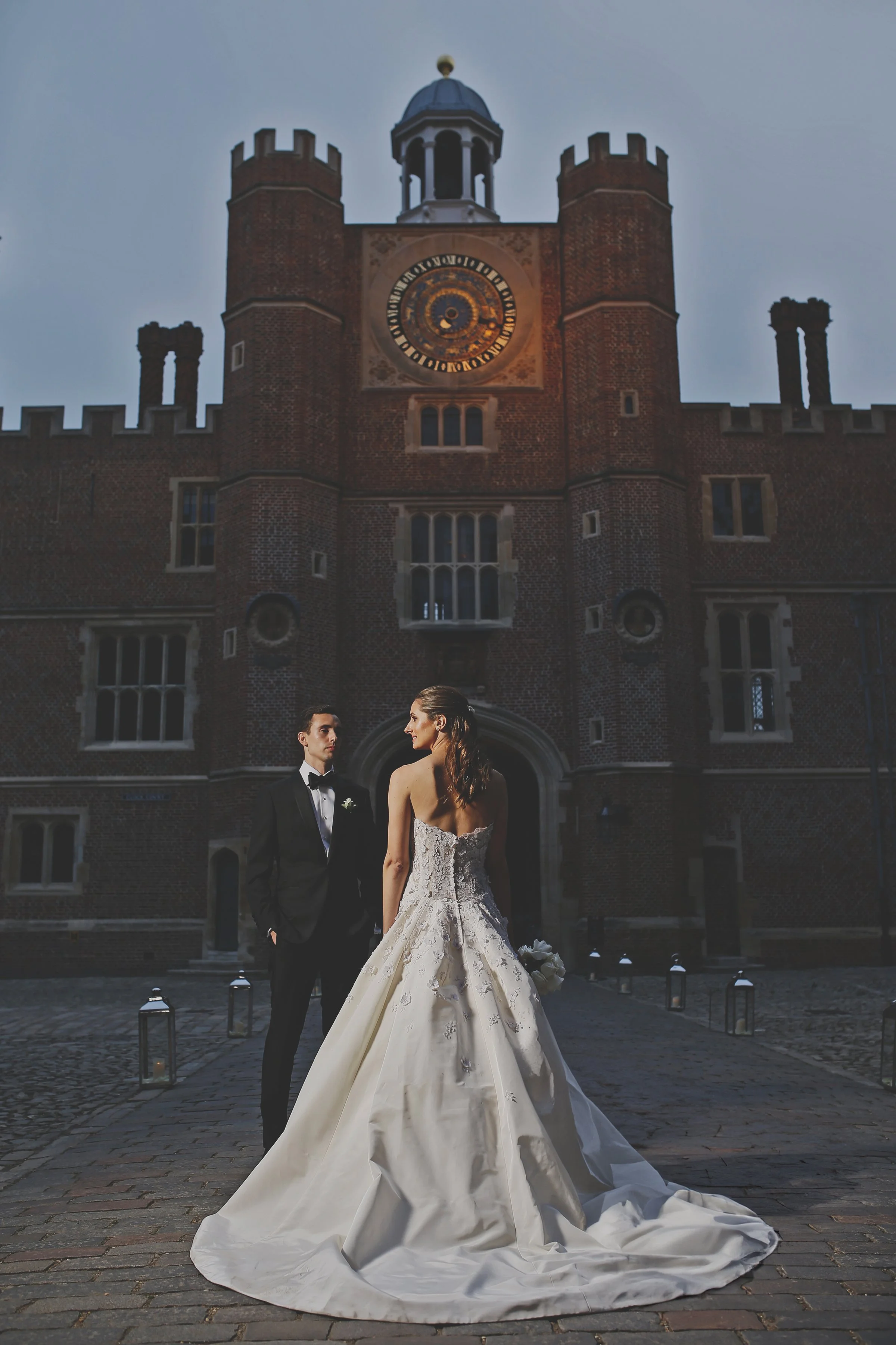 A bride and groom stand outside a historic building with a large clock tower. The bride is wearing a white gown and holding flowers. The groom is in a black suit. The building features brick walls and arched windows.