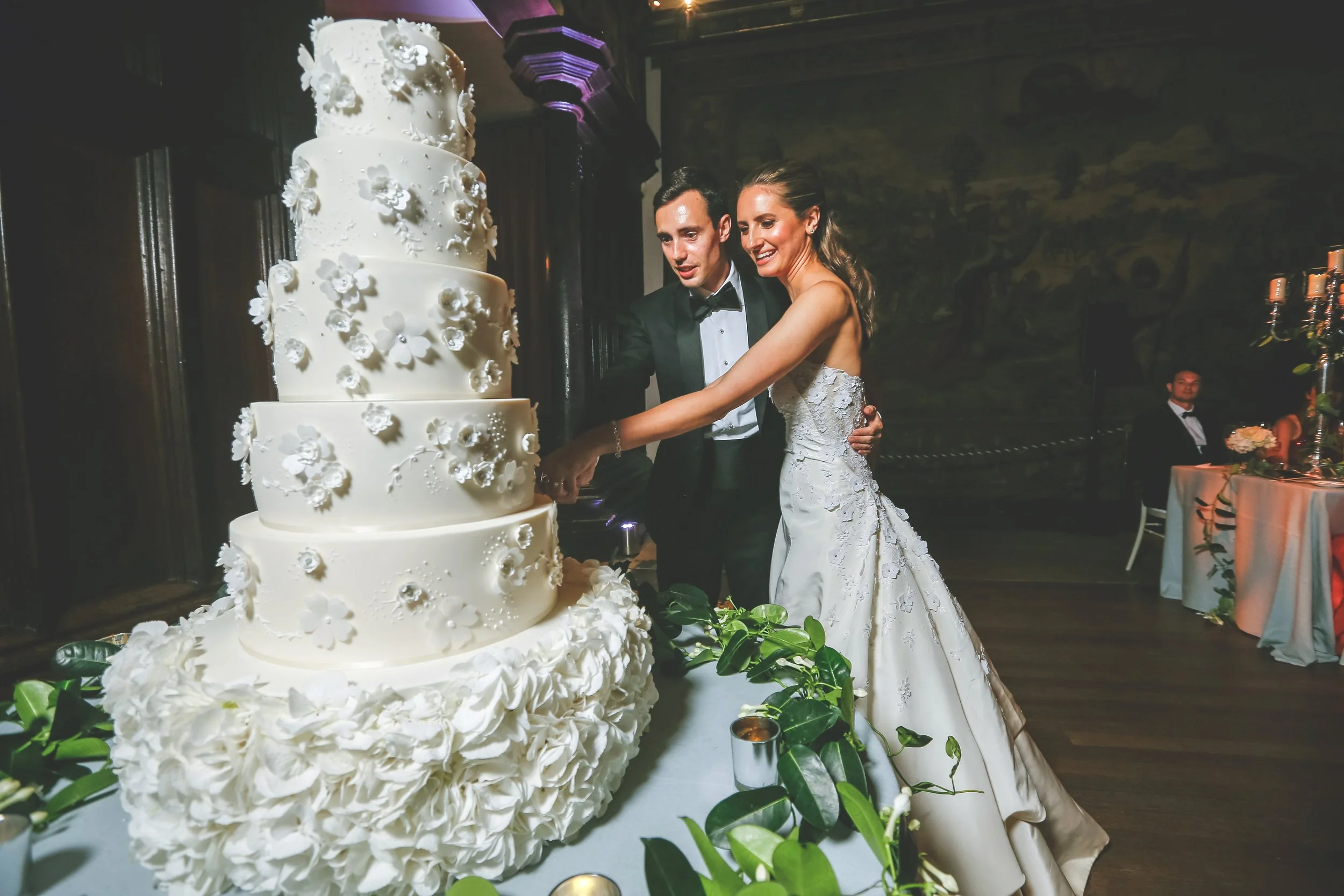 Bride and groom cutting a multi-tiered white wedding cake decorated with flowers, surrounded by greenery and candles.