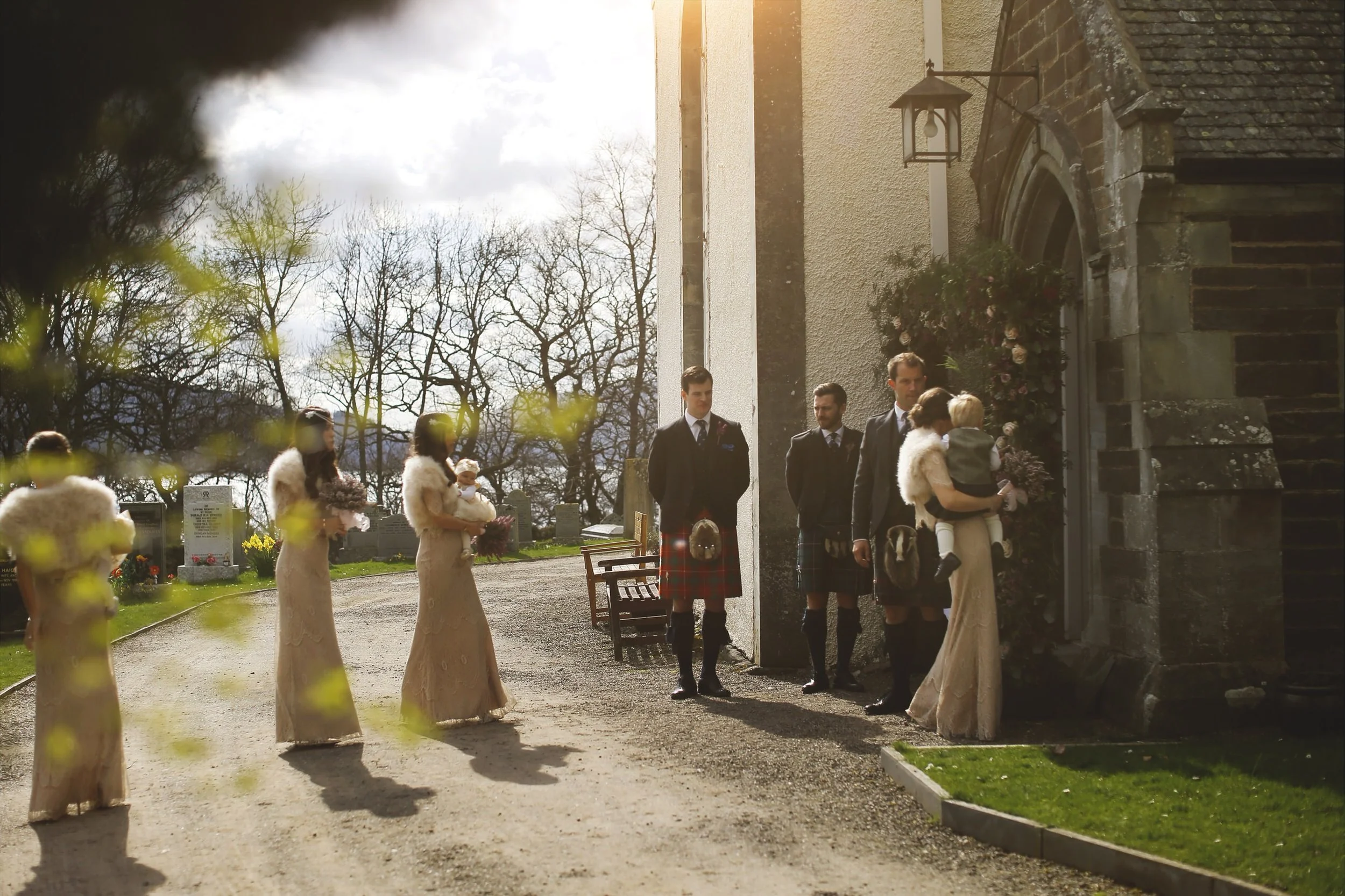 A wedding scene outside a church with four men in kilts and women in light-colored dresses holding children, on a sunny day with bare trees in the background.