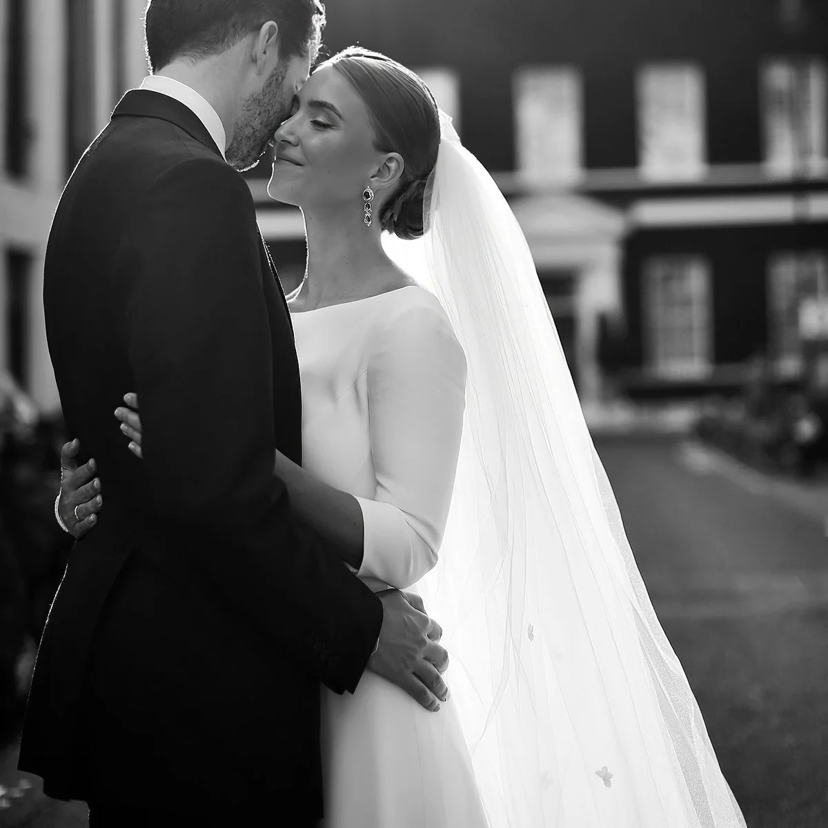 A black and white photo of a bride and groom embracing on their wedding day. The bride is wearing a long veil and a simple white dress, while the groom is in a suit. They are standing outside with a blurred building in the background.