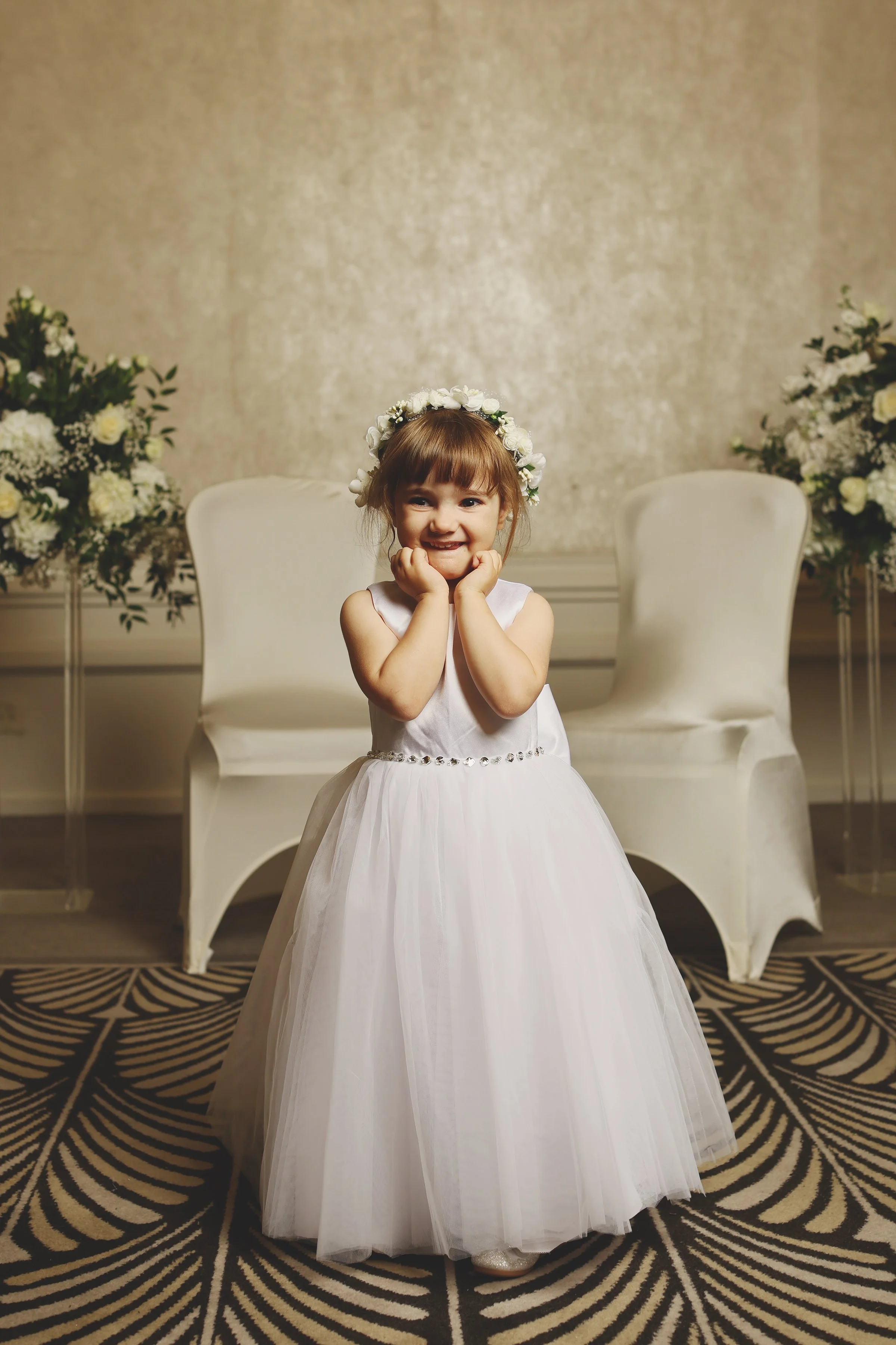 Smiling young girl in white dress and floral crown, standing in decorated room with white chairs and floral arrangements.