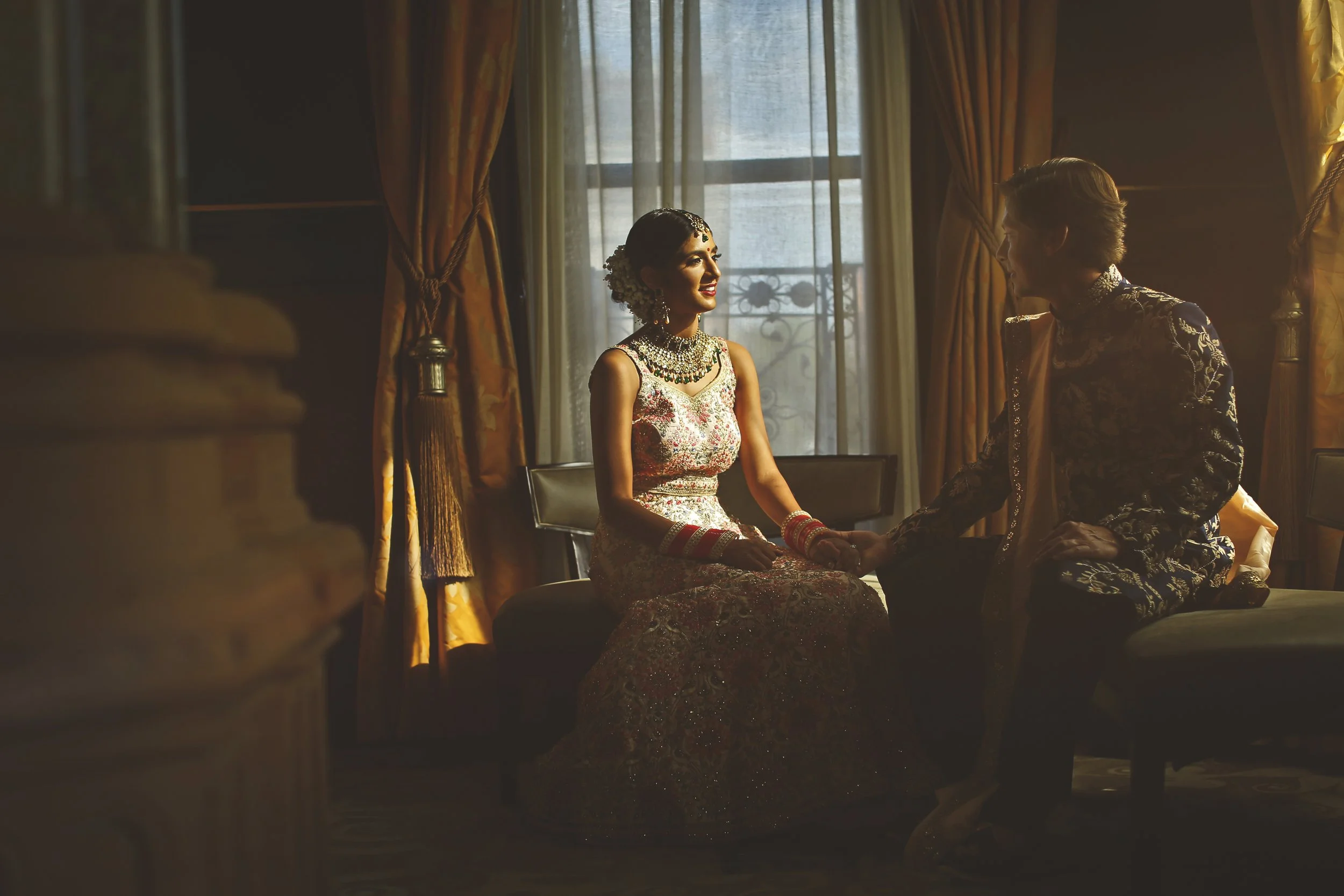 Bride and groom in traditional attire sitting together in a dimly lit room with ornate curtains.