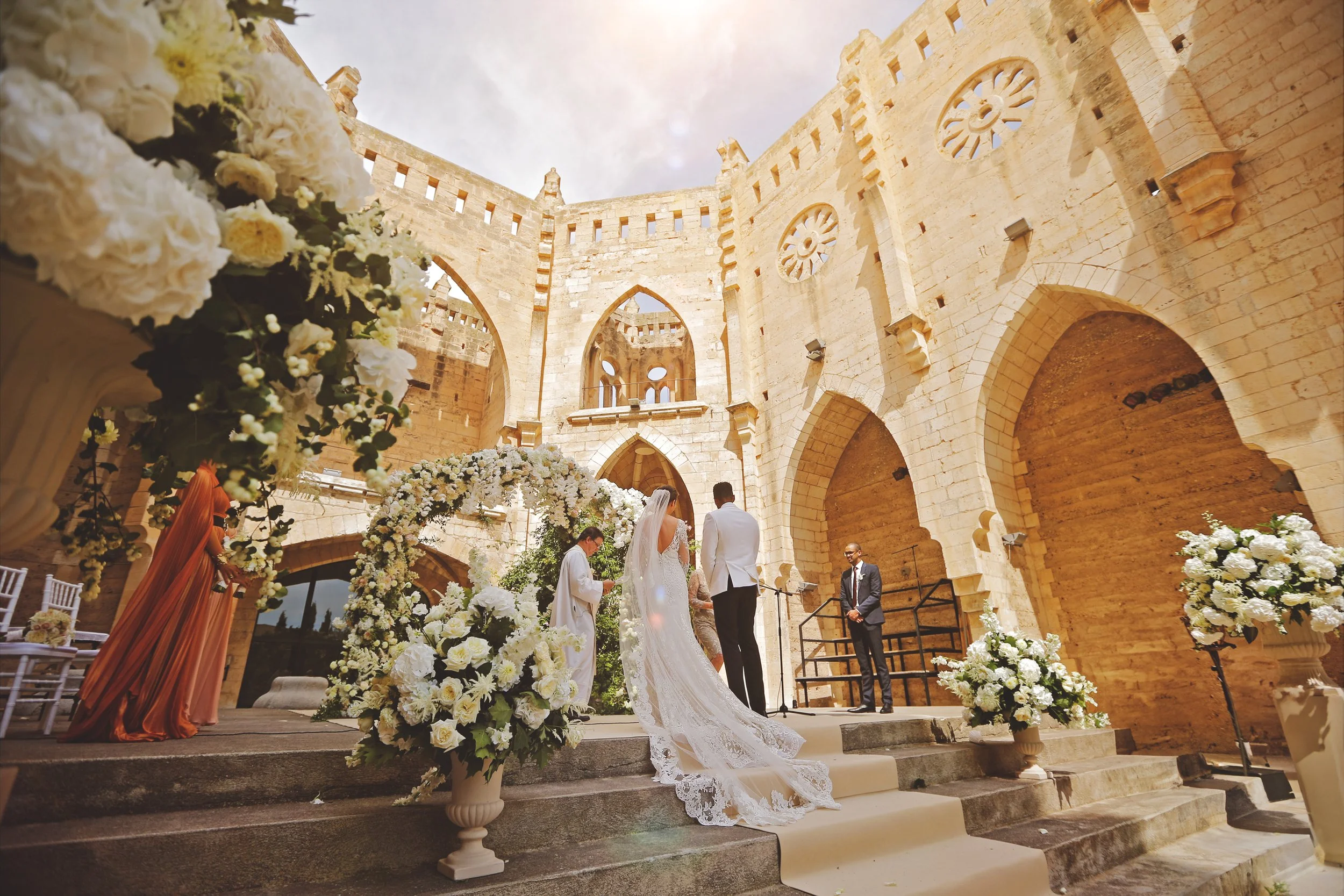 Bride and groom standing at the altar of an outdoor wedding ceremony, surrounded by floral arrangements and guests, with a historic building as the backdrop.