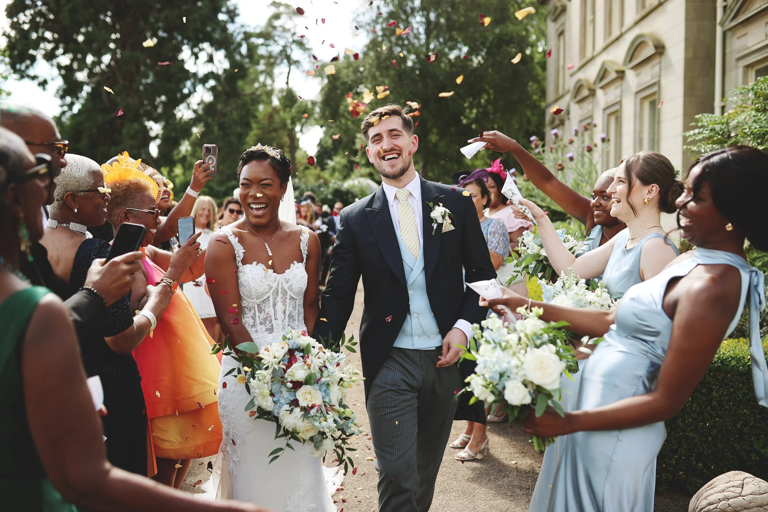 A bride and groom walk through guests throwing flower petals and taking photos outside in a garden setting.