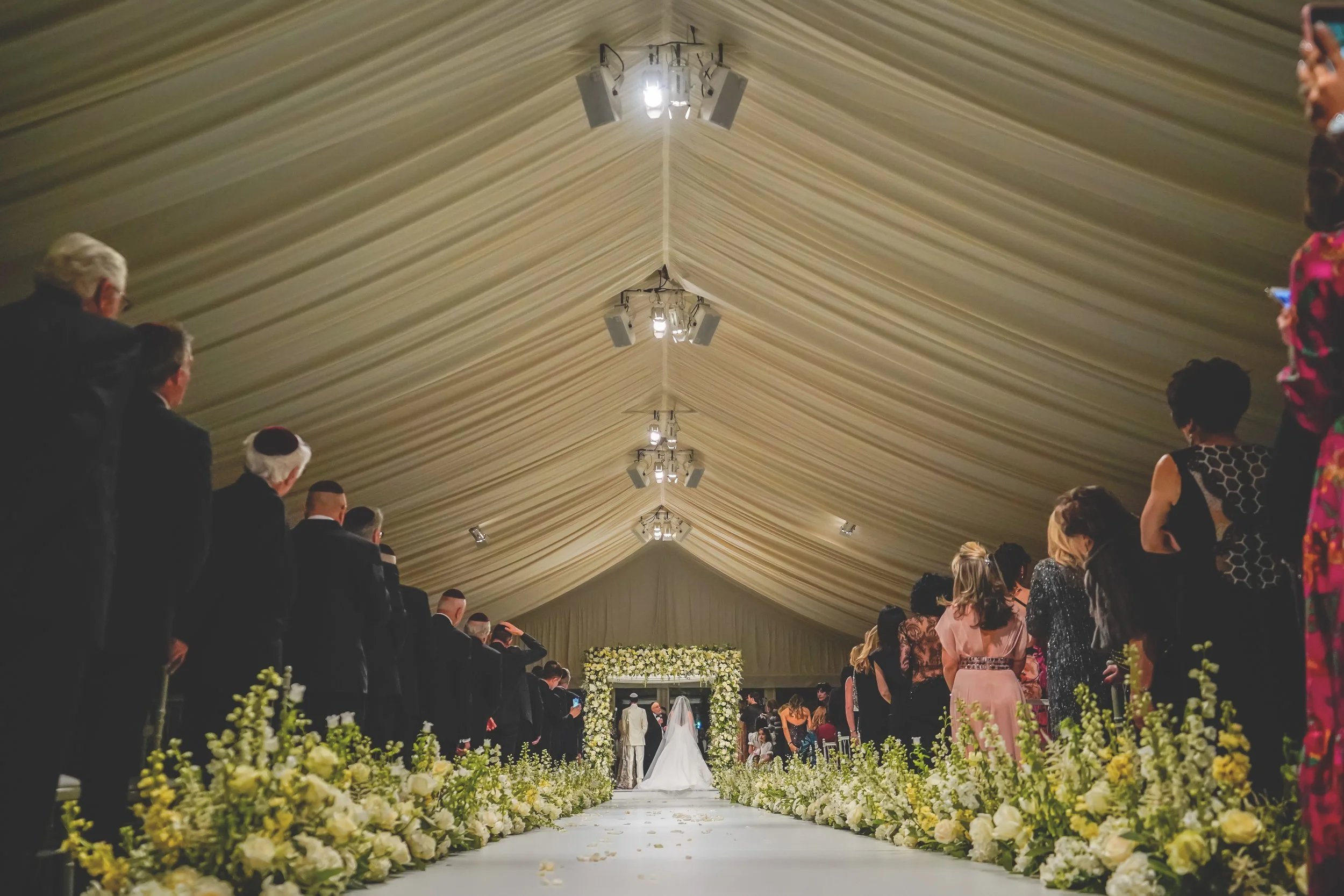 Jewish wedding ceremony in a decorated tent with guests and floral aisle.