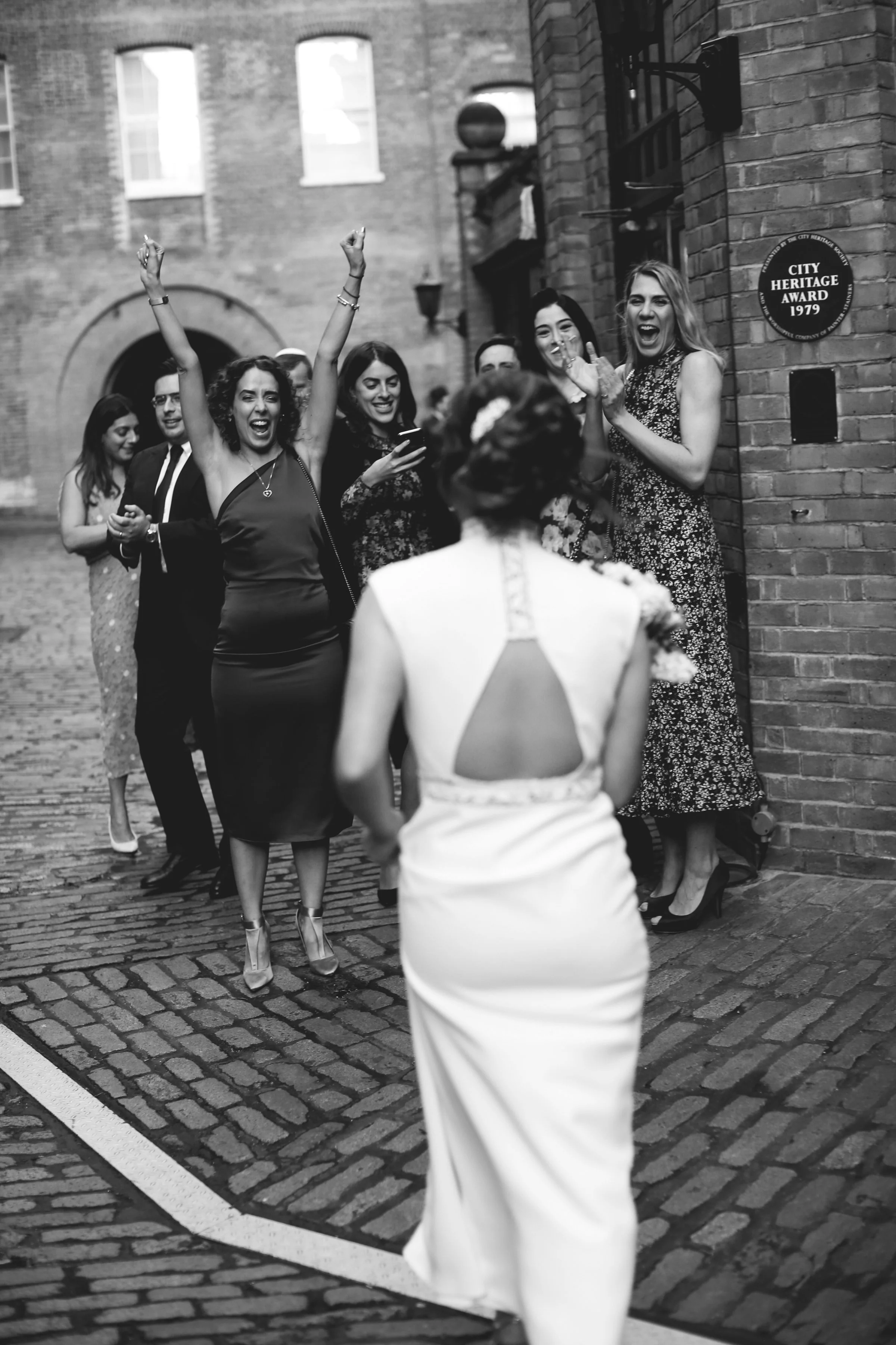 Bride in white dress approaching cheering group on cobblestone street, black and white photo