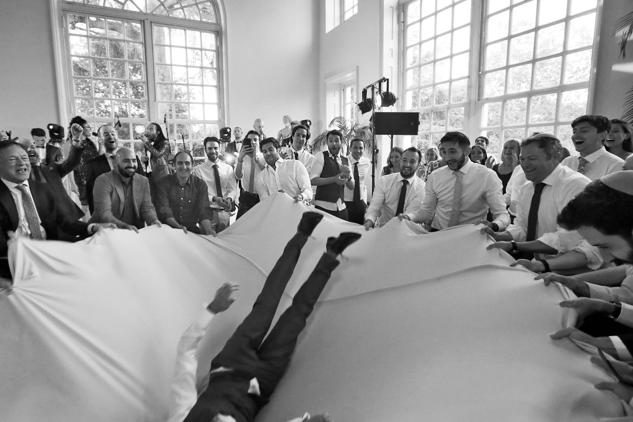 A group of people at a celebration holding a cloth, tossing a person into the air indoors.