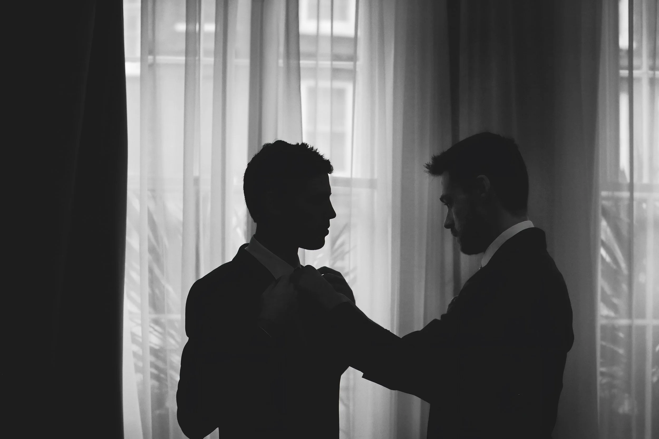 Silhouette of two men in suits, one adjusting the other's tie, standing in front of sheer curtains.