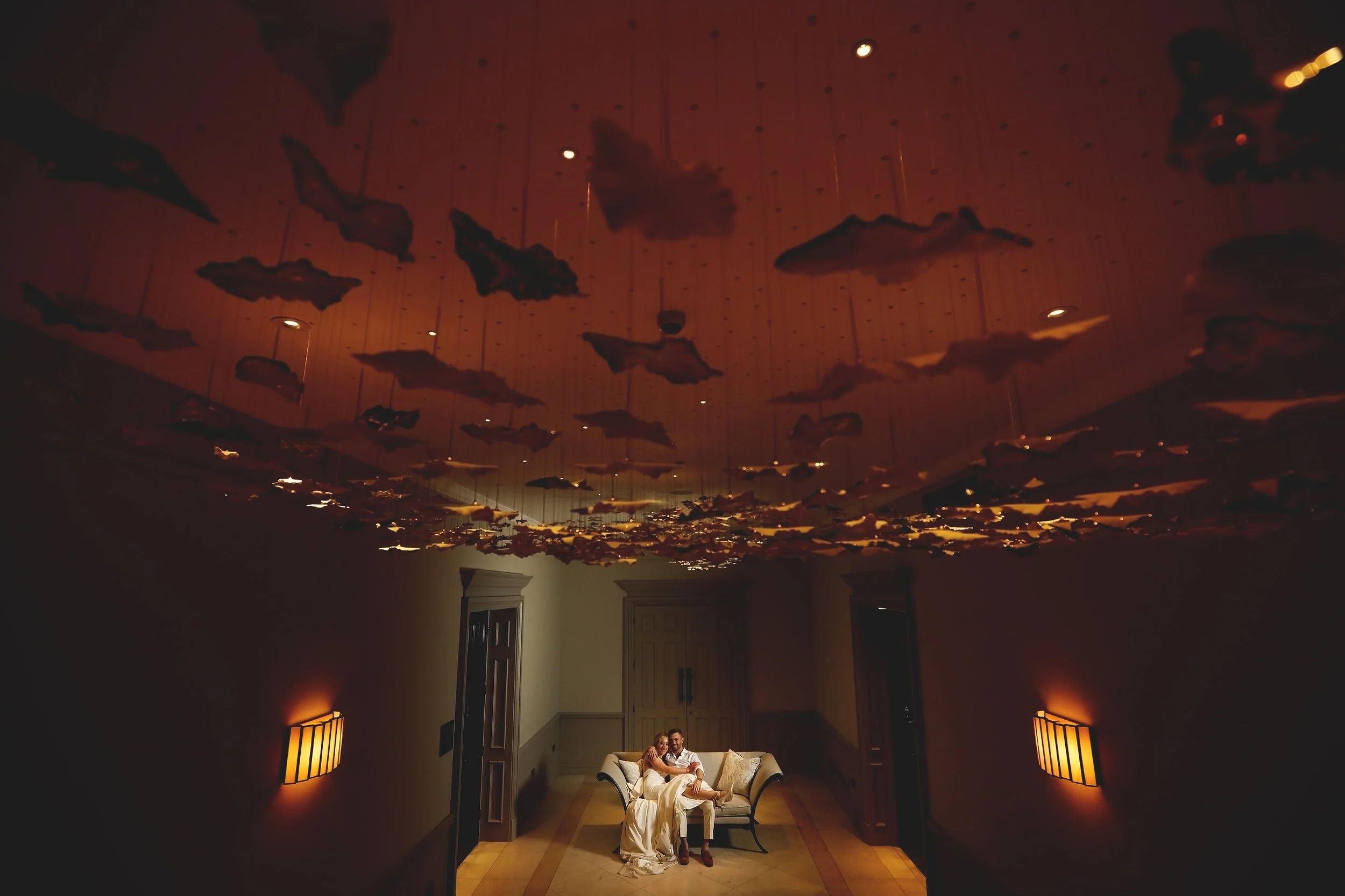 A couple seated on a sofa under a ceiling with artistic decorations resembling flying shapes. The room is dimly lit with warm lighting from wall sconces.