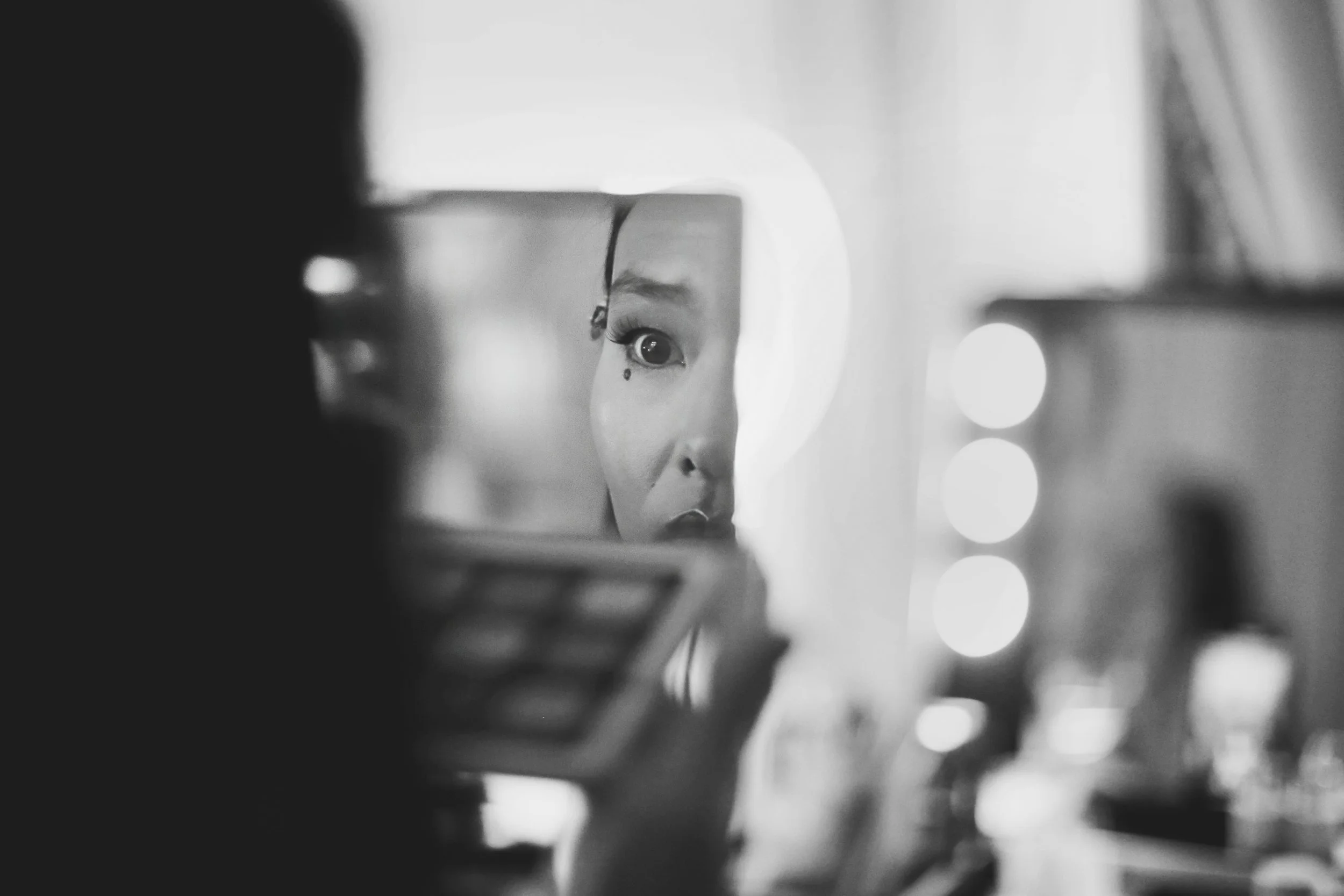 Black and white image of a person applying makeup while looking in a mirror, focusing on their eyes and an eyeshadow palette.