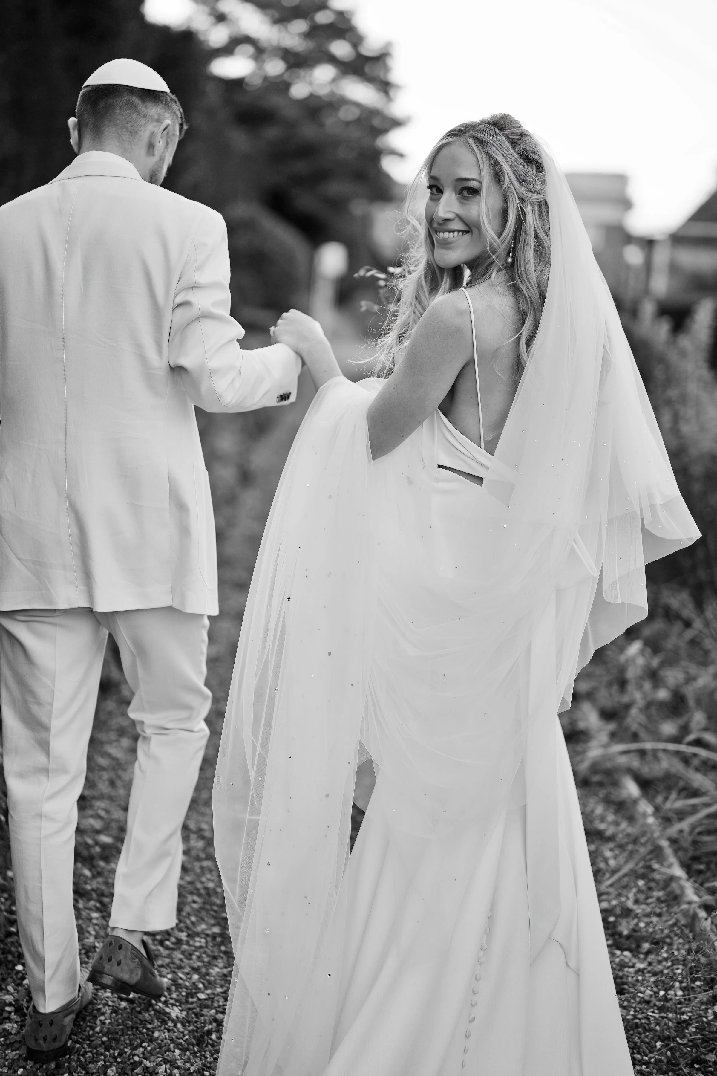 Bride smiling while holding hands with a man, both dressed in formal white attire, outside on a path; black and white photo.