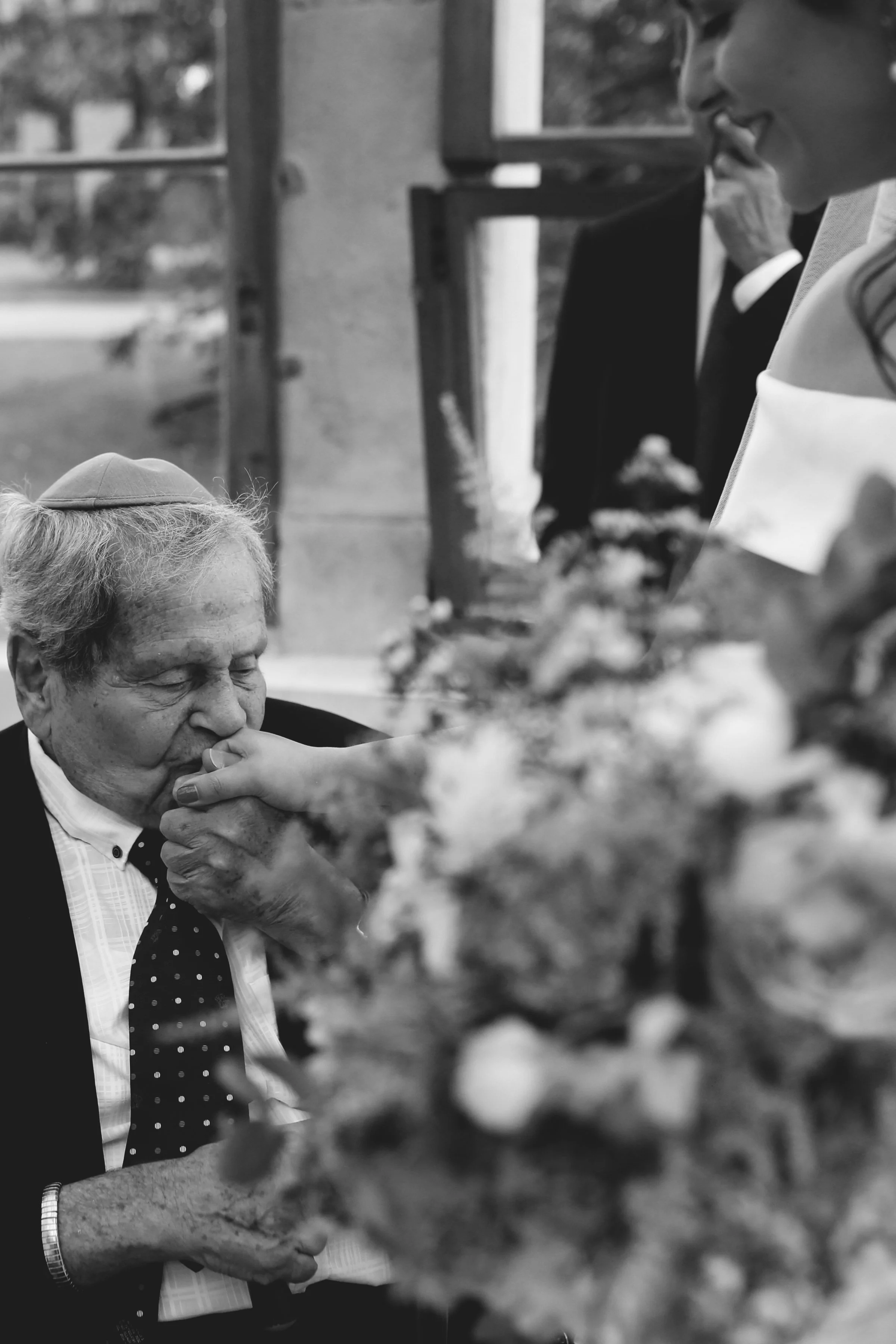 Elderly man wearing a kippah kissing a bride's hand, with wedding flowers in the foreground.