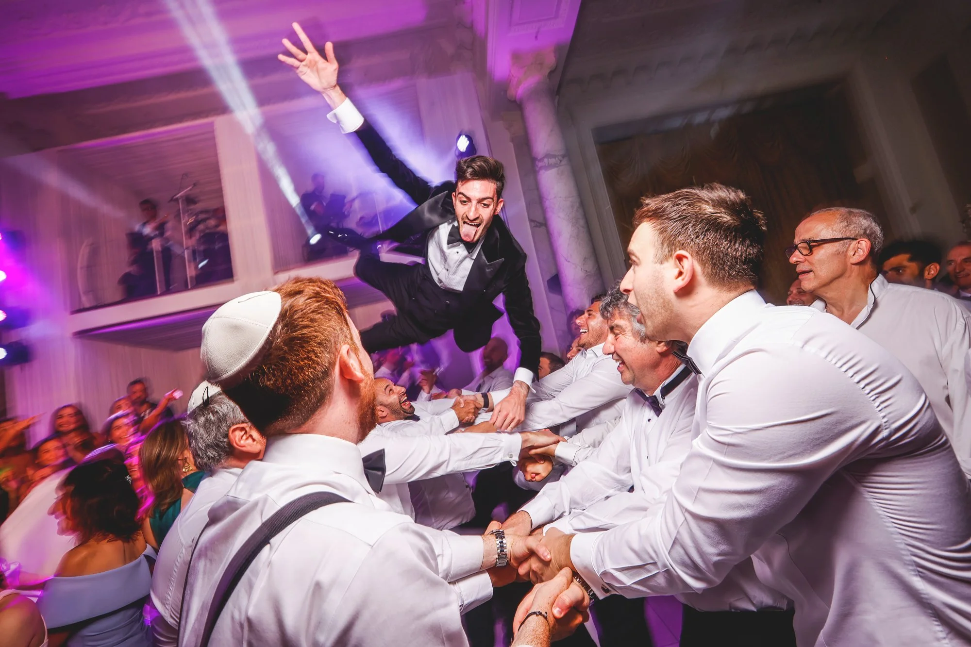 Group of people celebrating at a party, with a man being joyfully tossed in the air by others. Attendees are dressed in formal attire, and the atmosphere is festive with colorful lights.