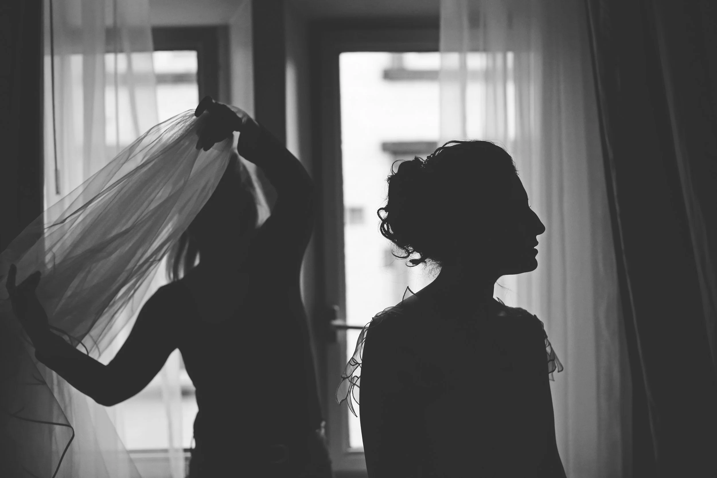 Silhouette of a bride standing indoors with another person adjusting a veil.