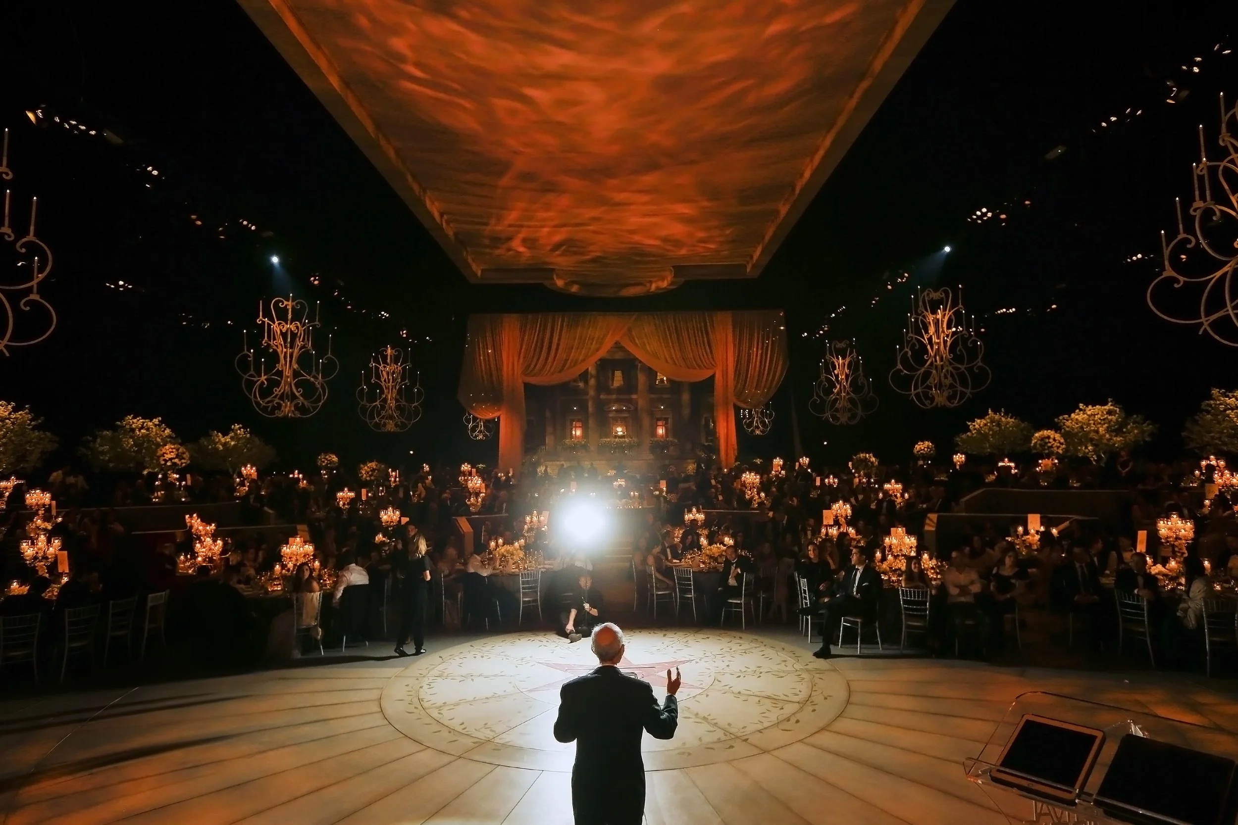 Elegant formal event with a speaker addressing an audience, featuring chandeliers, candlelit tables, and draped backdrops.