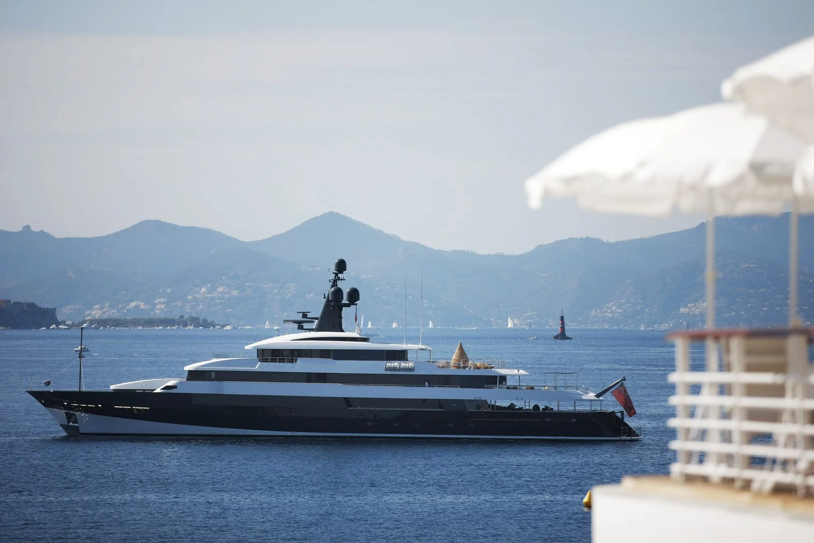 A large black and white yacht on calm blue water with a mountainous landscape in the background. There are white umbrellas partially visible in the foreground on the right side.