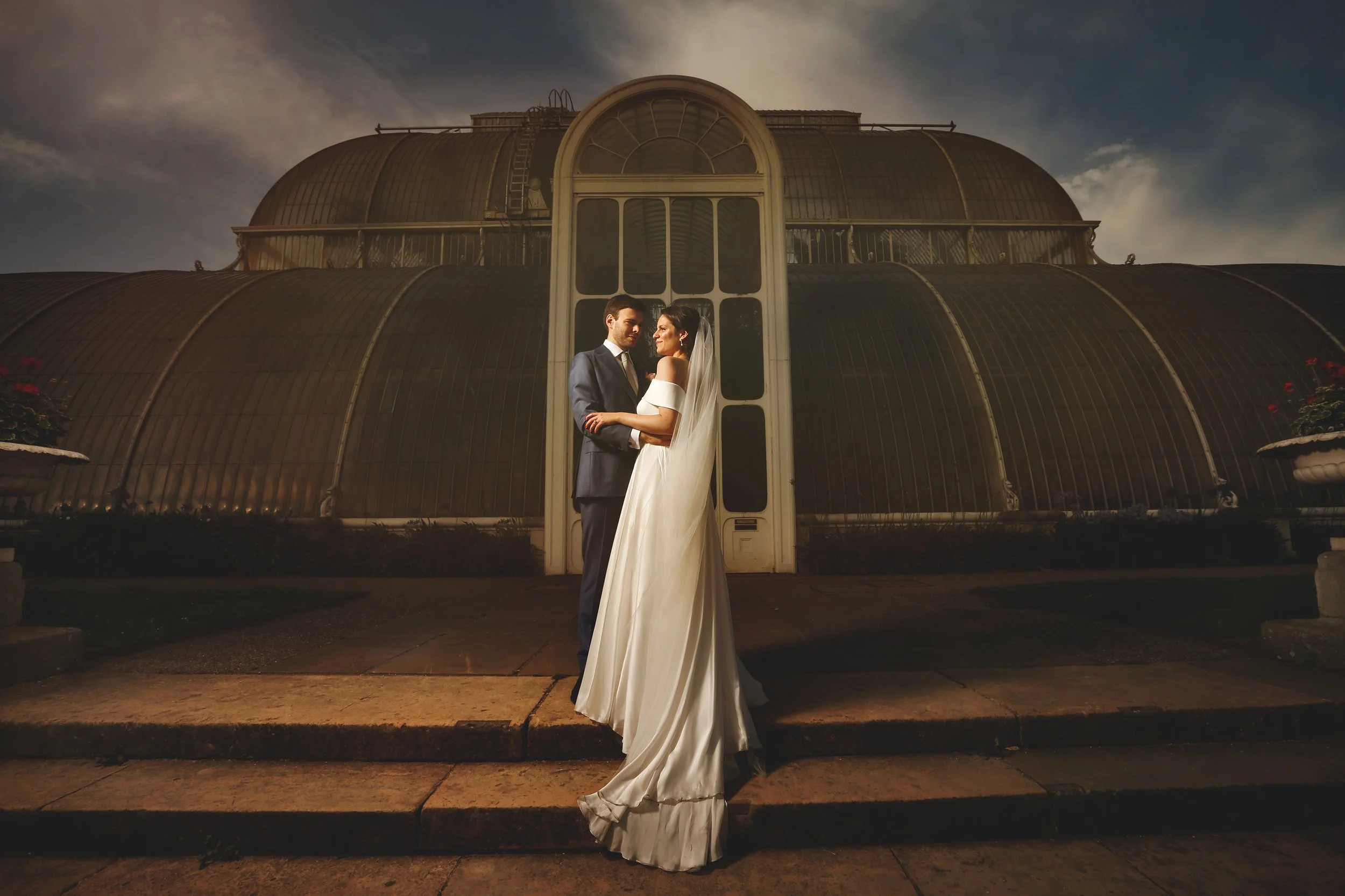 Bride and groom standing in front of a large greenhouse structure, with the bride in a white gown and veil and the groom in a dark suit, both looking at each other.