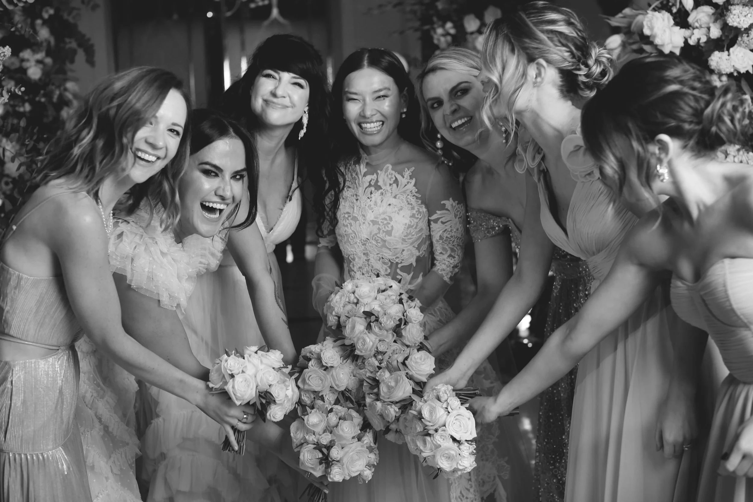Bridal party smiling, holding bouquets, wearing formal dresses, black and white photo.