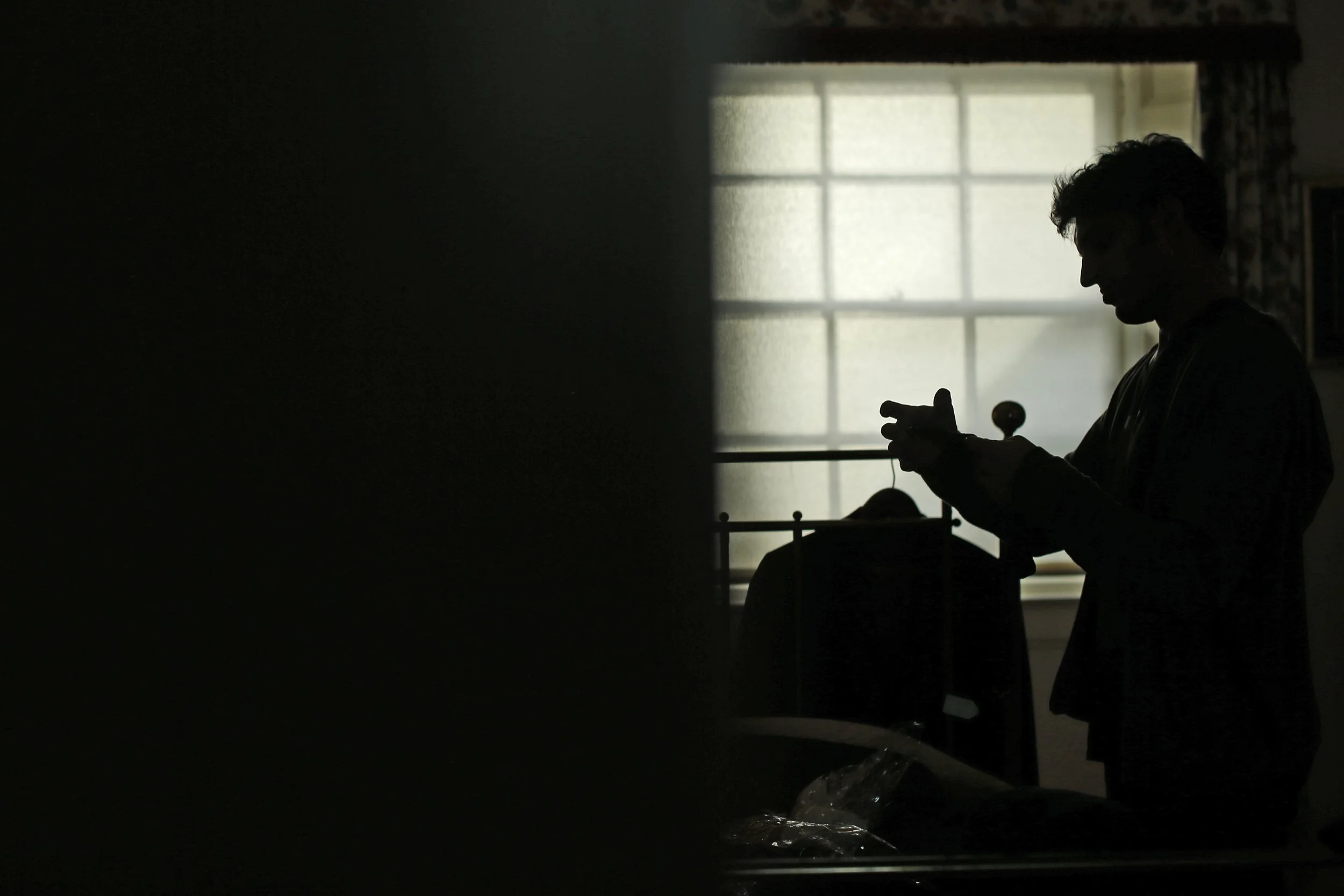 A dark silhouette of a person standing in front of a window, with soft backlighting illuminating the outline of their hair and face, holding an object in their hands, possibly a phone or small device, in a room with a bed and a nightstand nearby.
