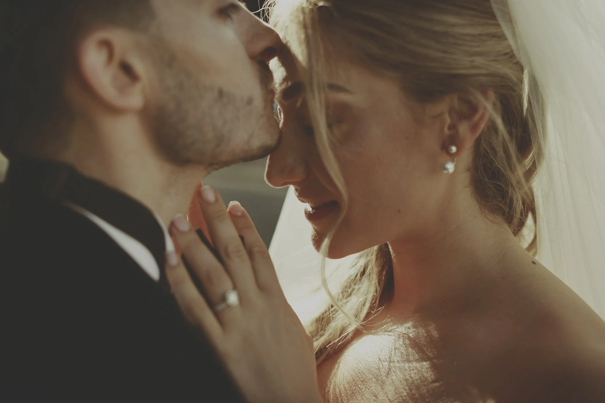 A groom kissing a bride's forehead, close-up, showing engagement ring and veil.