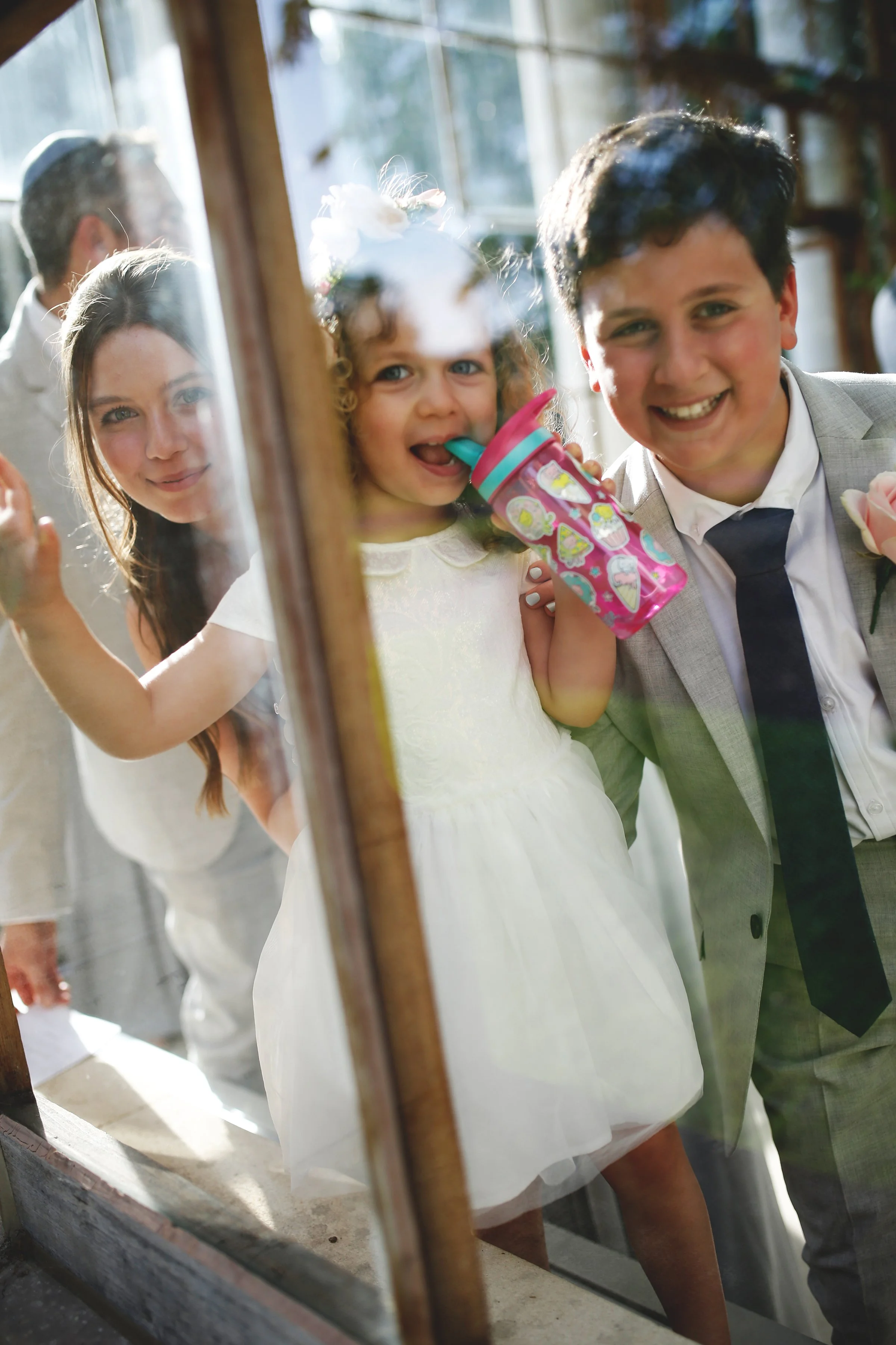 Three smiling children, two boys and a girl, looking through a glass window. The girl is holding a pink sippy cup. The boy in a suit has a rose on his lapel.