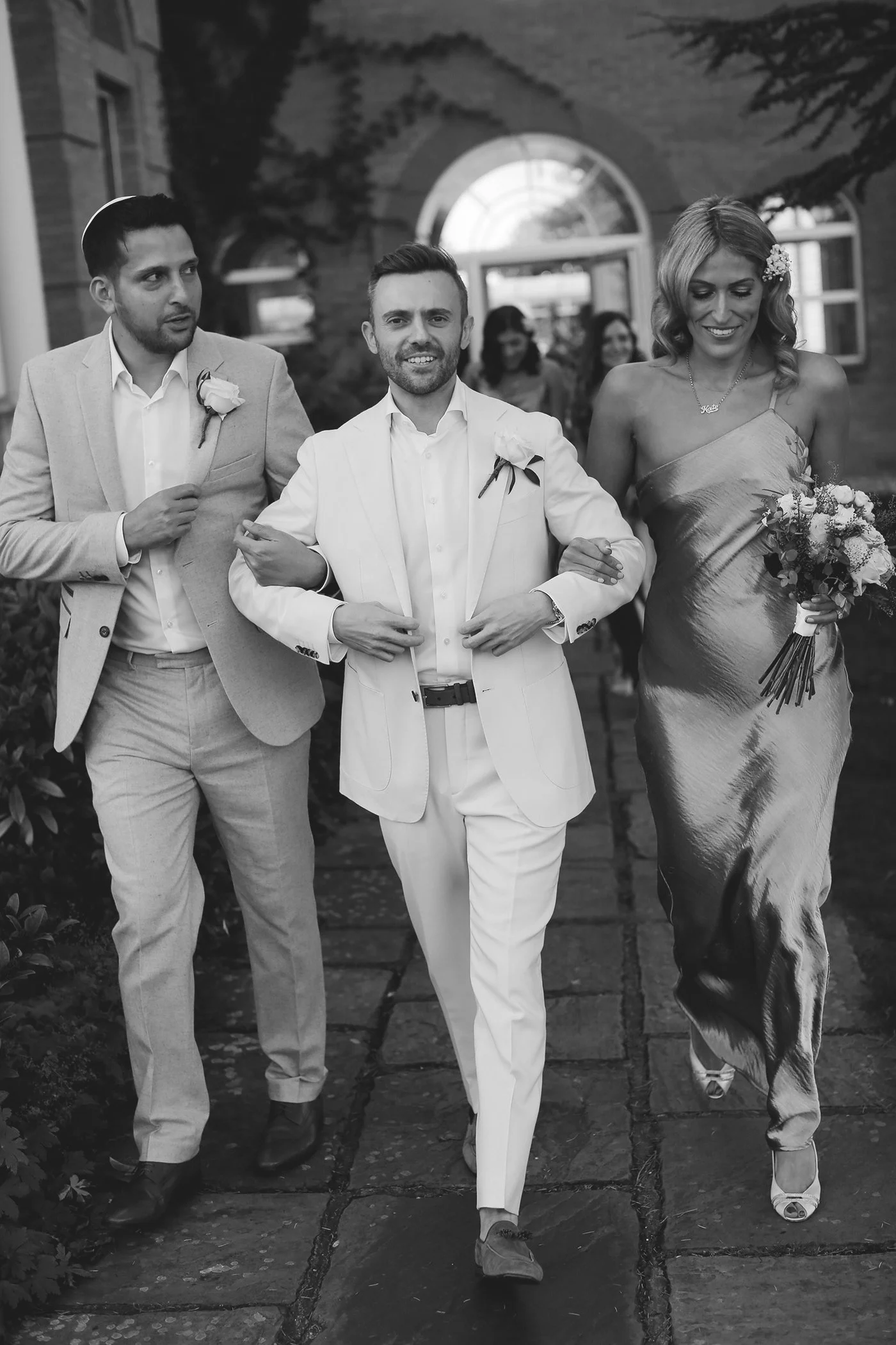 Three people walking at a wedding, two men in suits and a woman in a dress holding flowers.