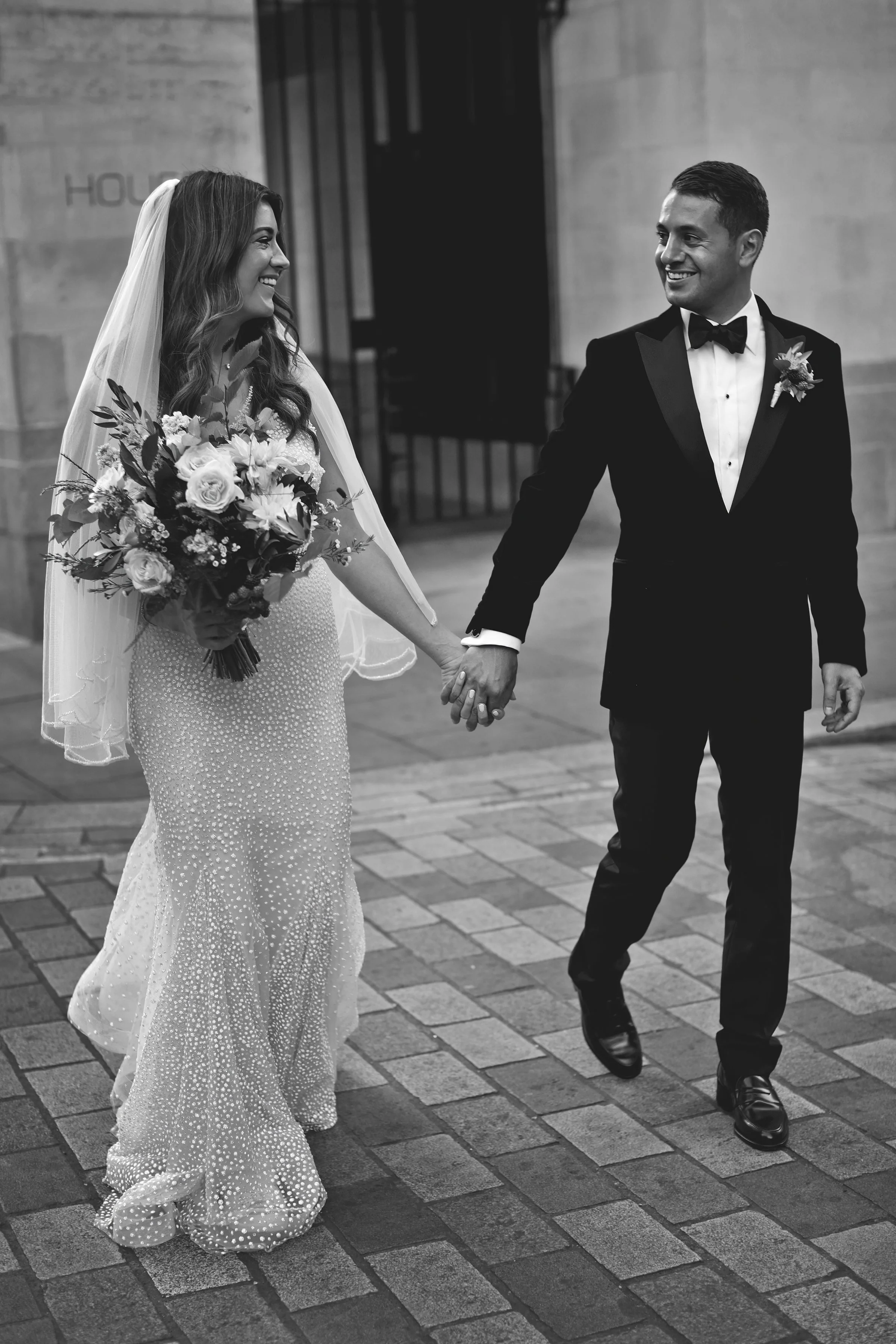 A bride and groom holding hands and smiling at each other. The bride is wearing a long, embellished gown and veil, holding a bouquet. The groom is in a black tuxedo with a bow tie. The image is in black and white.