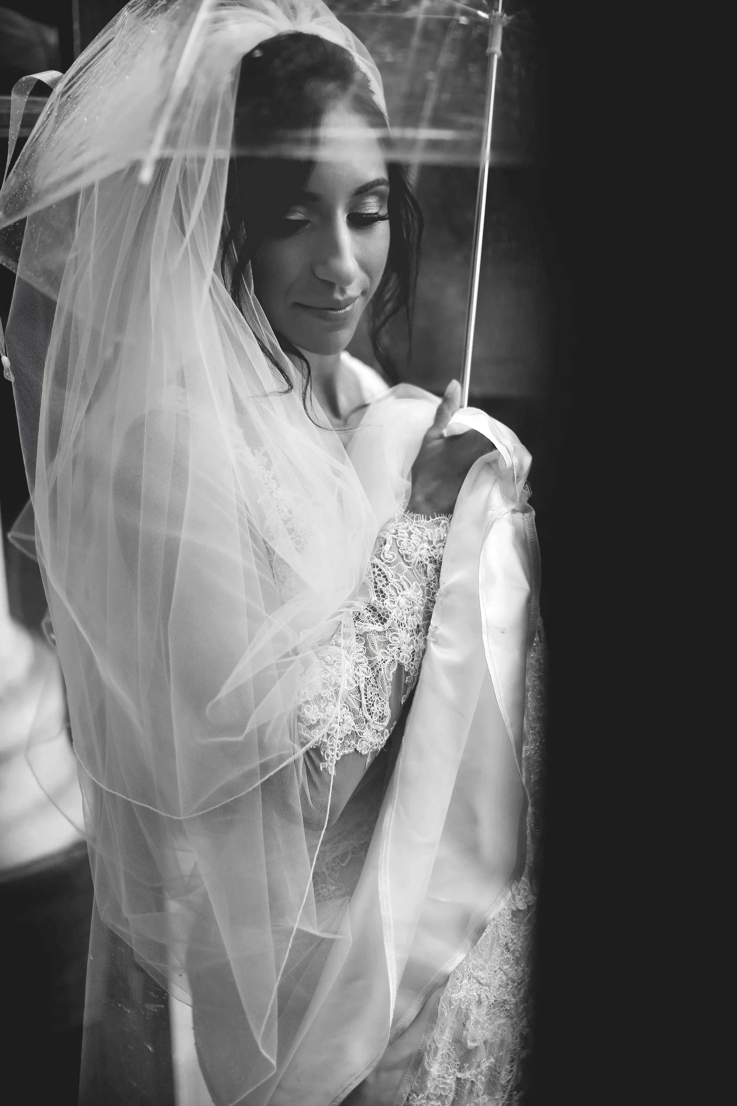 Bride holding a sheer umbrella, wearing a lace wedding dress and veil, captured in black and white.